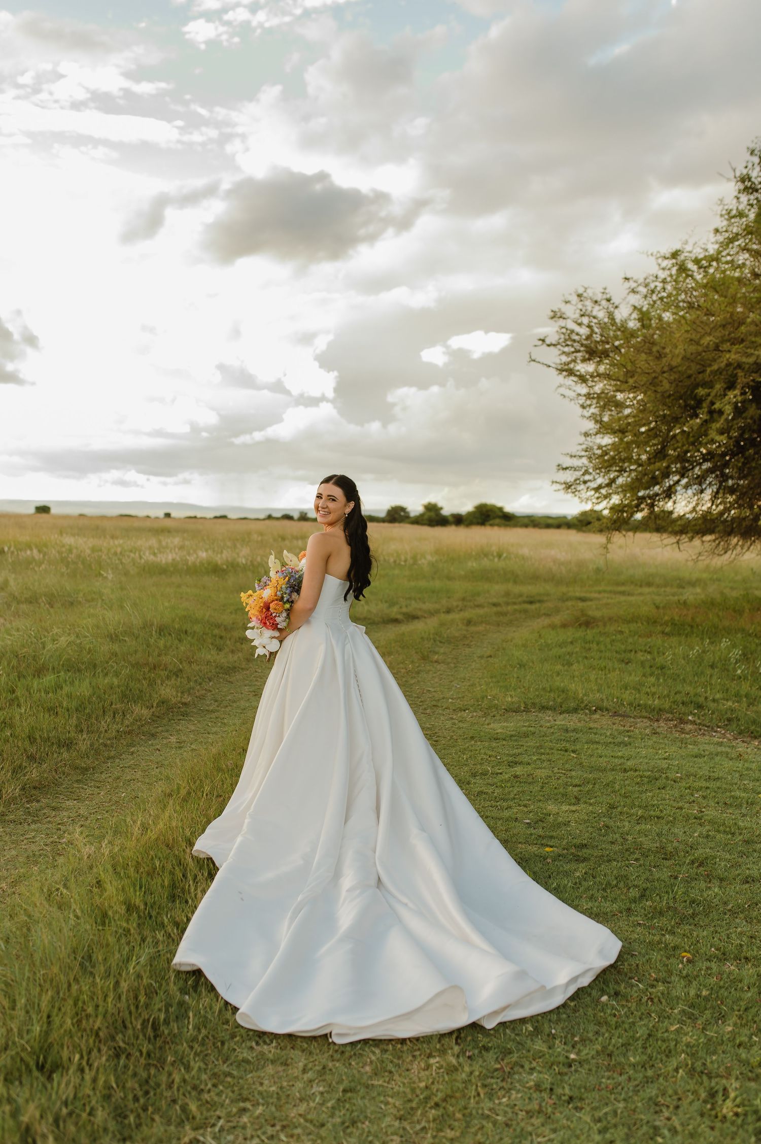 Bride in white strapless gown holding colorful bouquet standing in open green field.