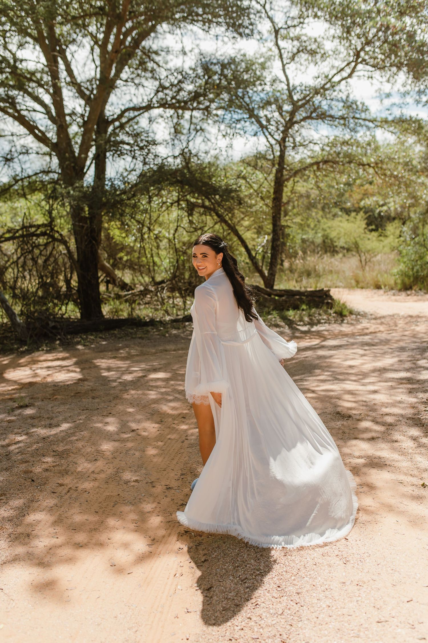 Bride in a flowing white bridal robe twirling on a sandy path surrounded by African bush trees.