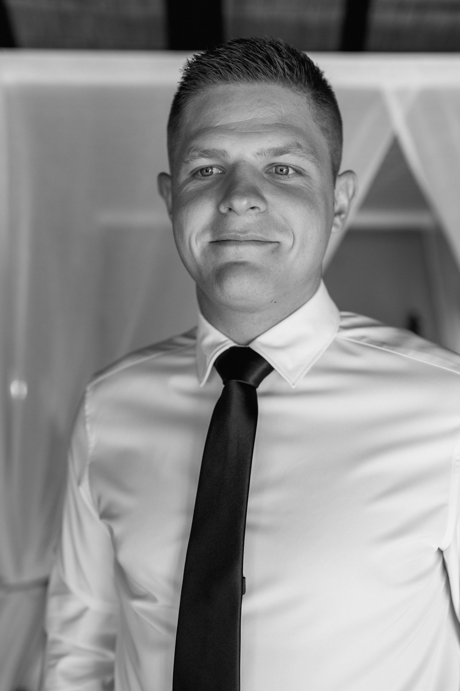 Black and white portrait of a groom in white dress shirt and black tie smiling confidently.