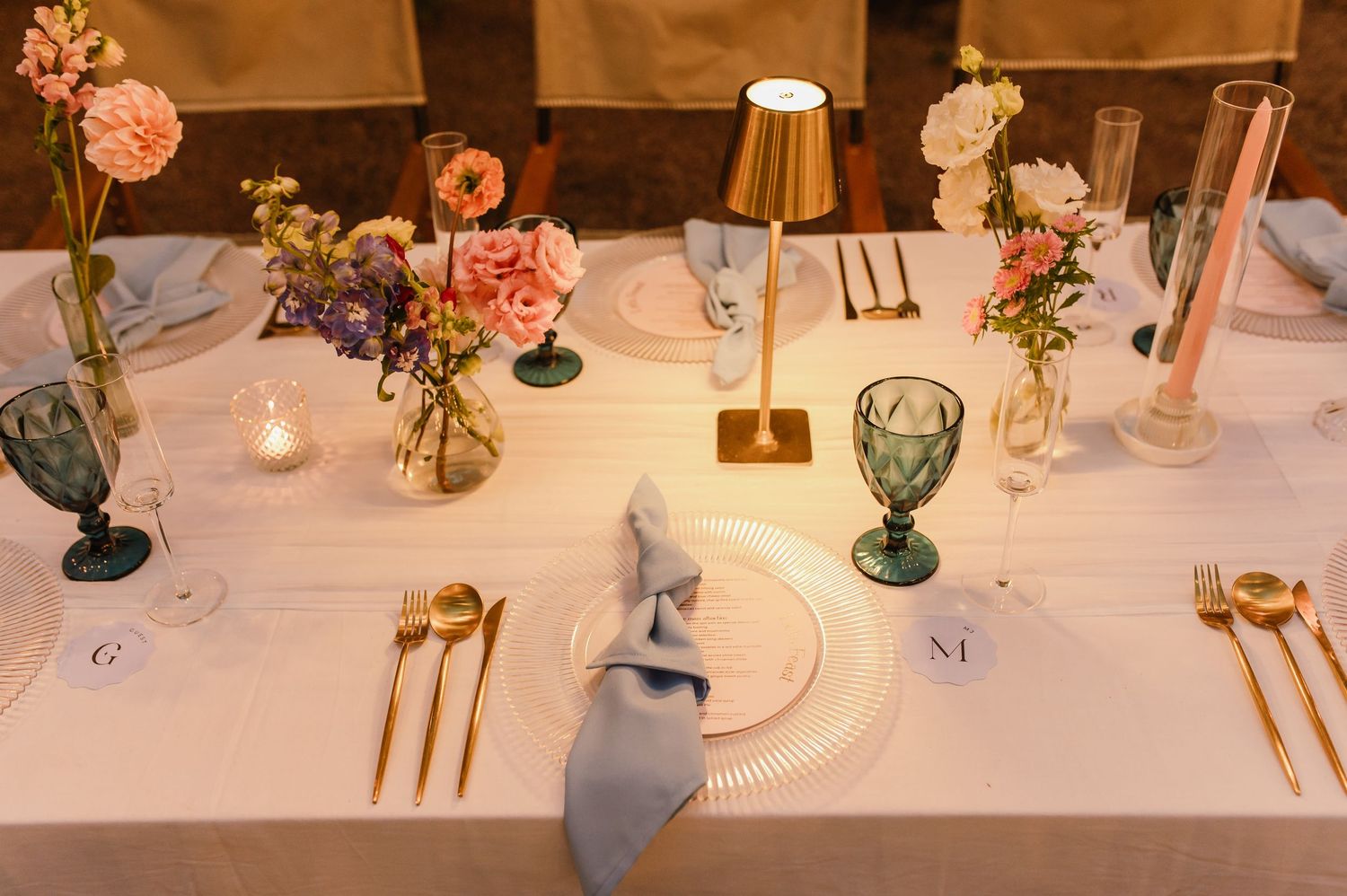 Elegant wedding reception table set with gold cutlery, teal glasses, pink florals, and small gold lamps.