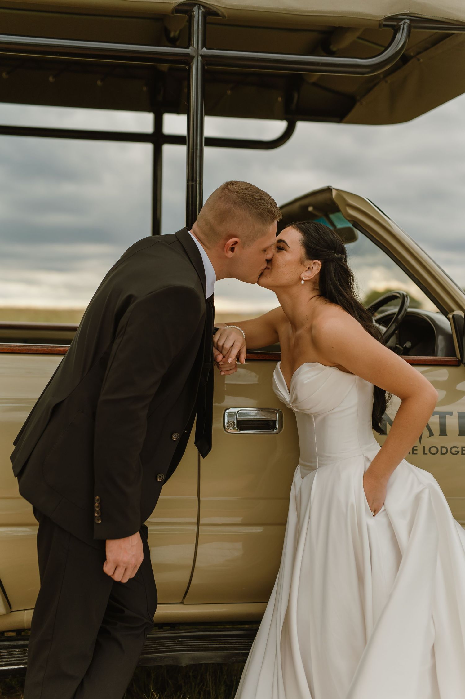 Bride and groom share romantic kiss leaning against vintage safari vehicle at outdoor wedding.