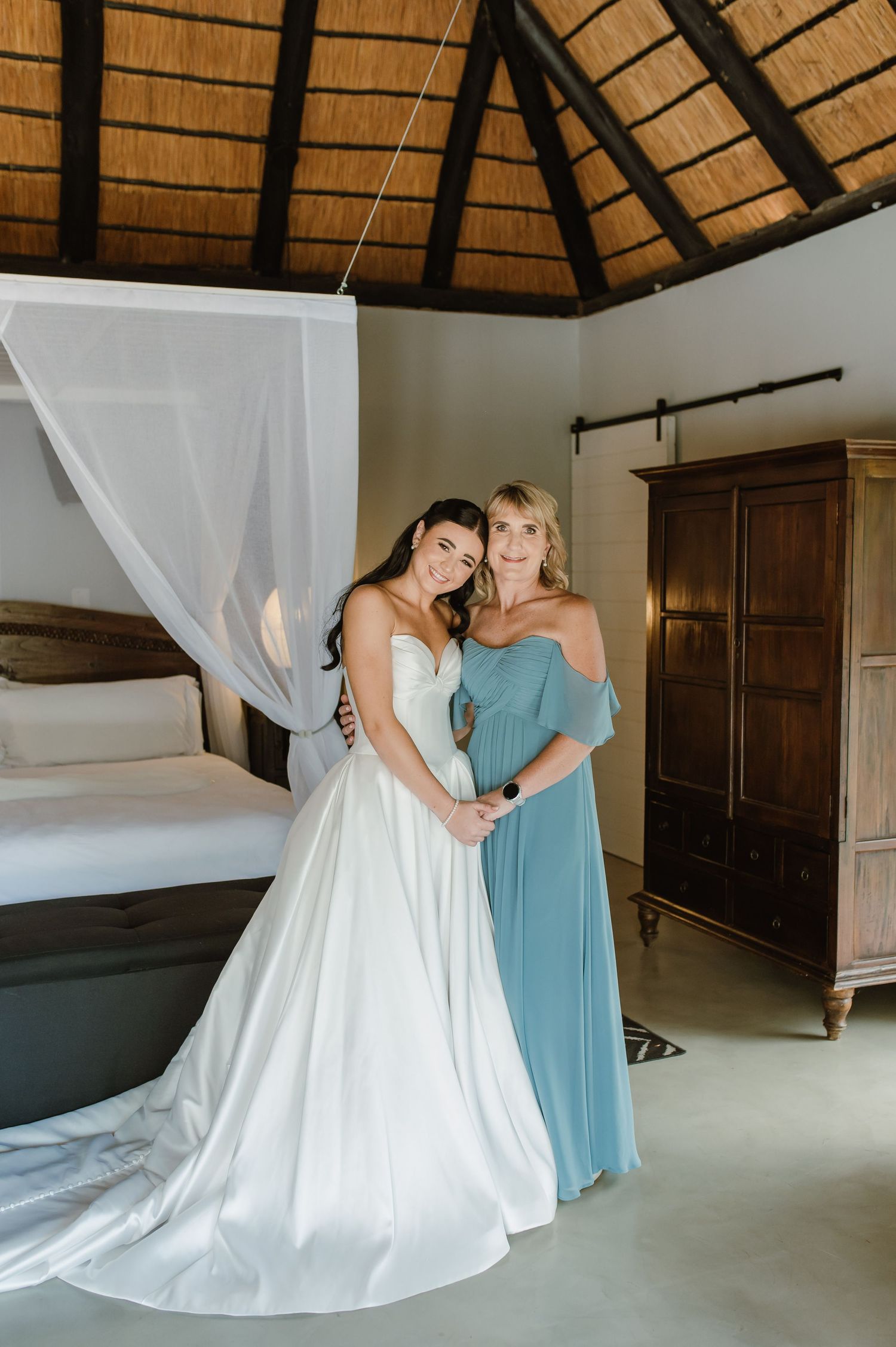 Bride in strapless white gown embraces woman in blue dress inside a thatched-roof wedding suite.