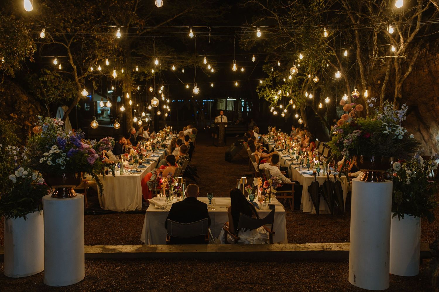 Elegant outdoor wedding reception with long tables, string lights, white pillar florals, and seated guests at night.
