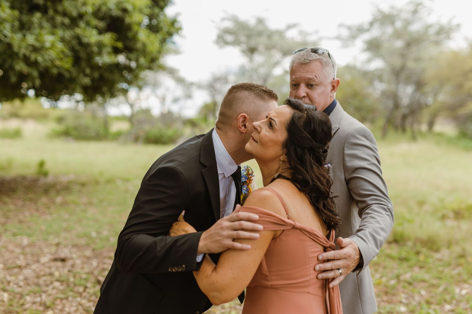 Two men kissing a smiling woman in a peach dress on the cheeks outdoors at a wedding celebration.