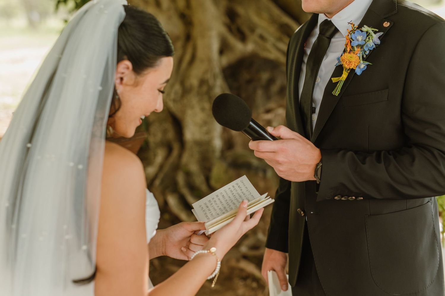 Bride reading wedding vows from small book while groom holds microphone during intimate outdoor wedding ceremony.