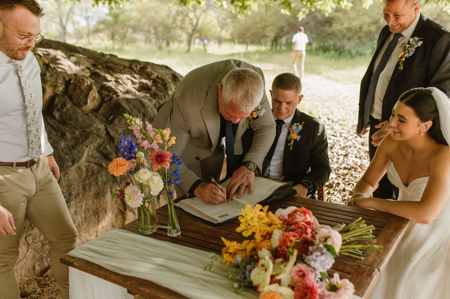 Wedding party signing marriage certificate at rustic wooden table decorated with vibrant wildflower bouquets.