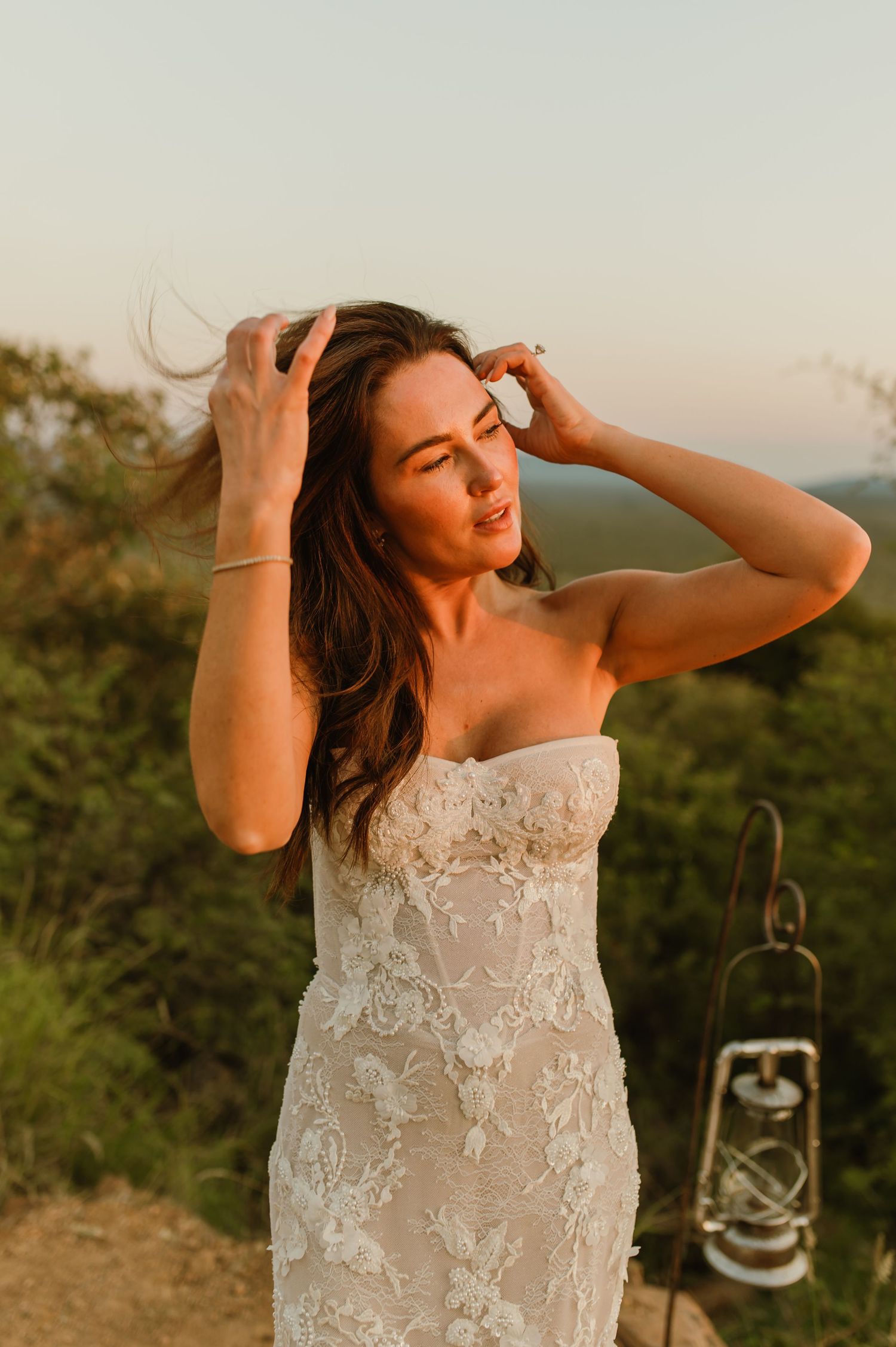 Bride in strapless lace wedding gown poses at golden hour with mountain views and vintage lantern backdrop.