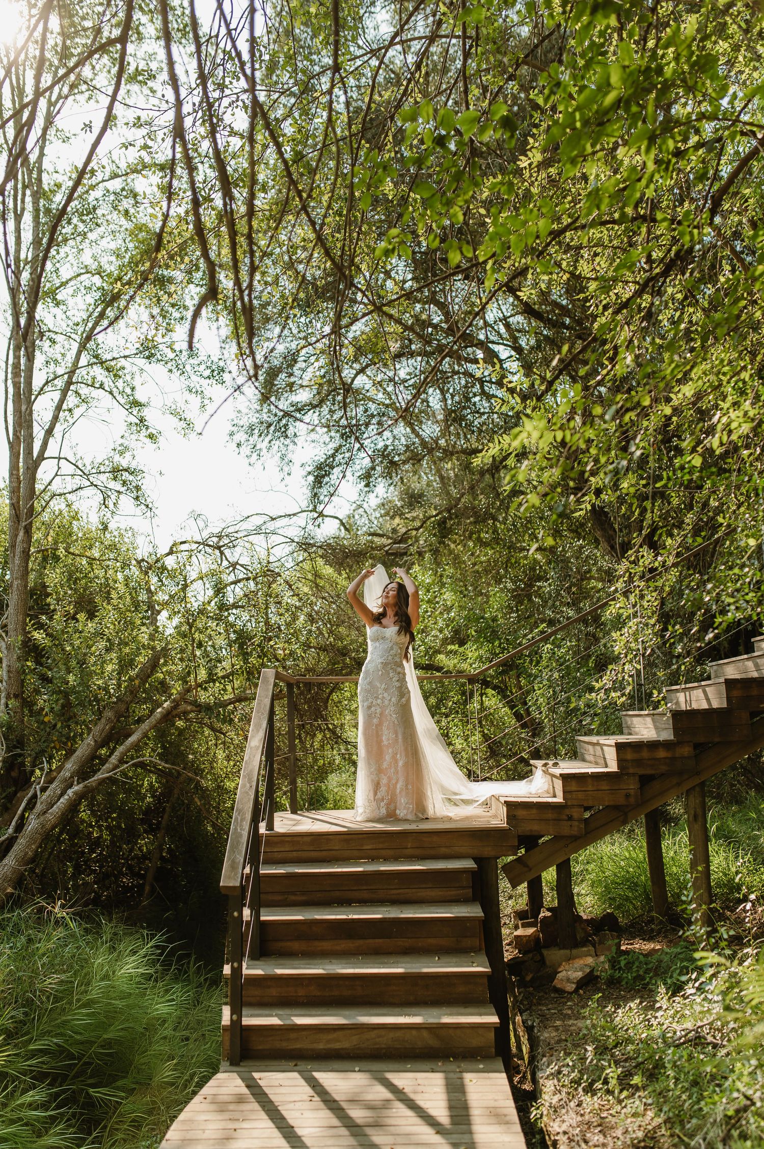 A bride in a lace wedding gown stands atop wooden stairs surrounded by lush green trees.