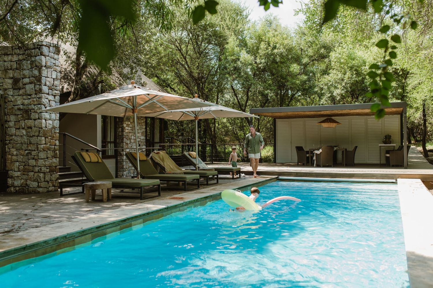 Family enjoying a luxury villa pool surrounded by lush trees with lounge chairs and umbrella.