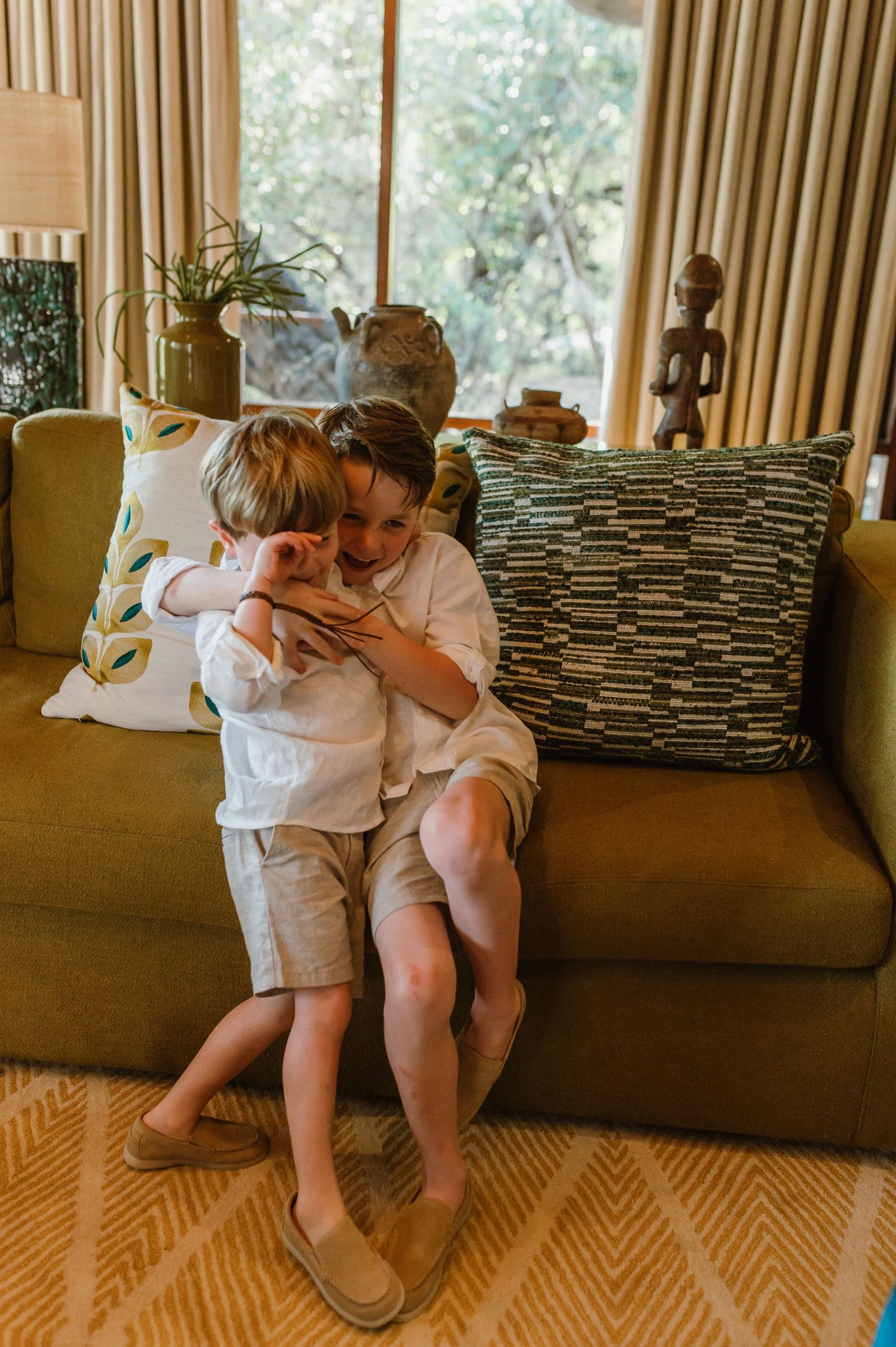 Two young boys share a warm hug on a mustard yellow sofa in a cozy living room.