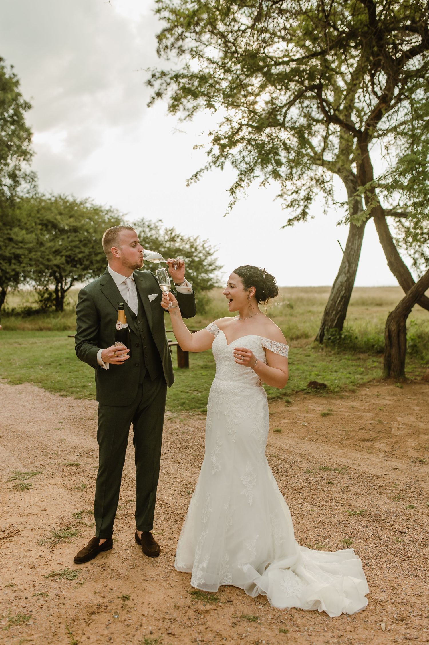 A couple in wedding attire shares a playful toast outdoors under trees in a rustic setting.