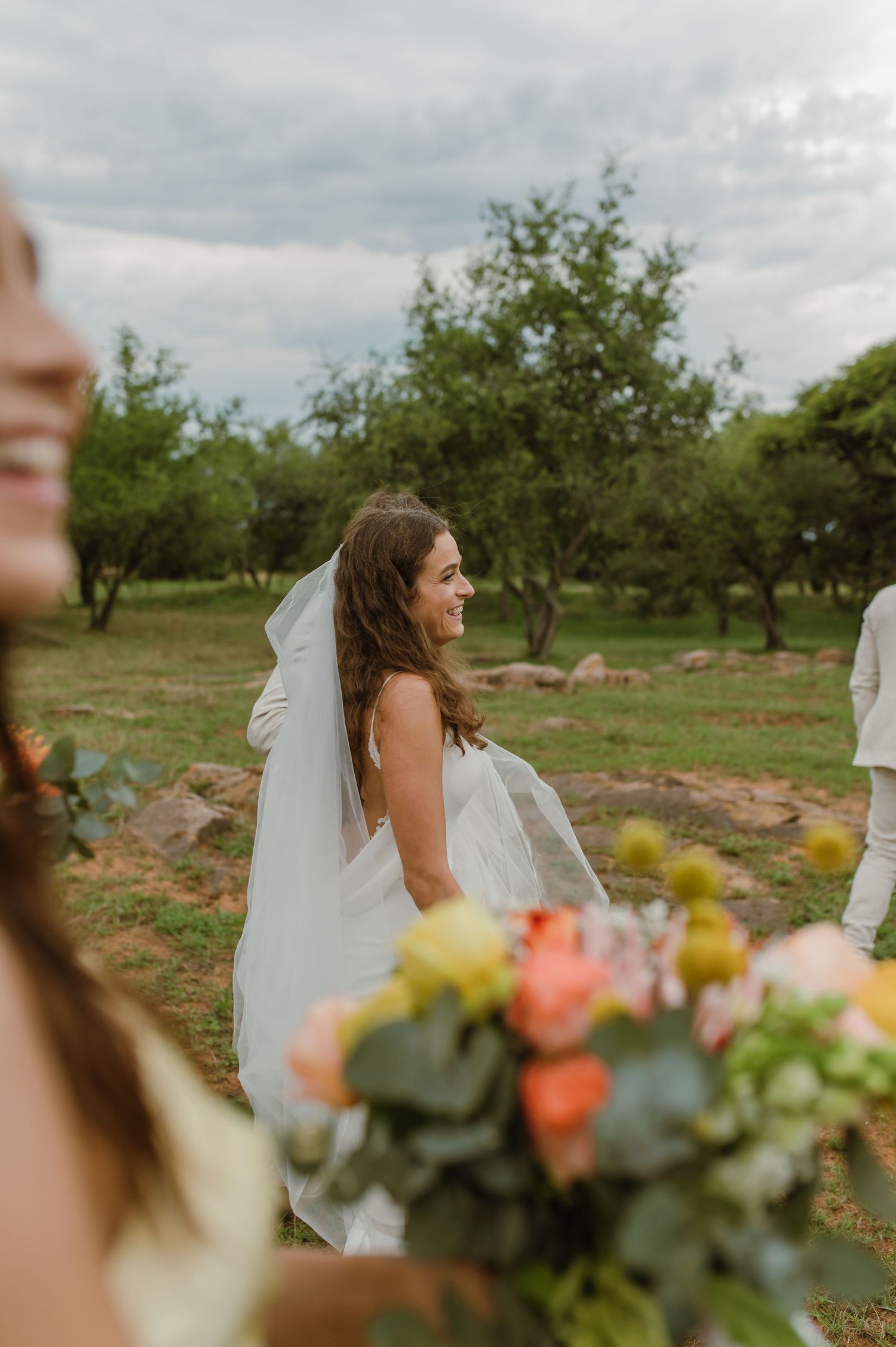 A wedding party poses in a natural outdoor setting with trees and flowers in soft lighting.