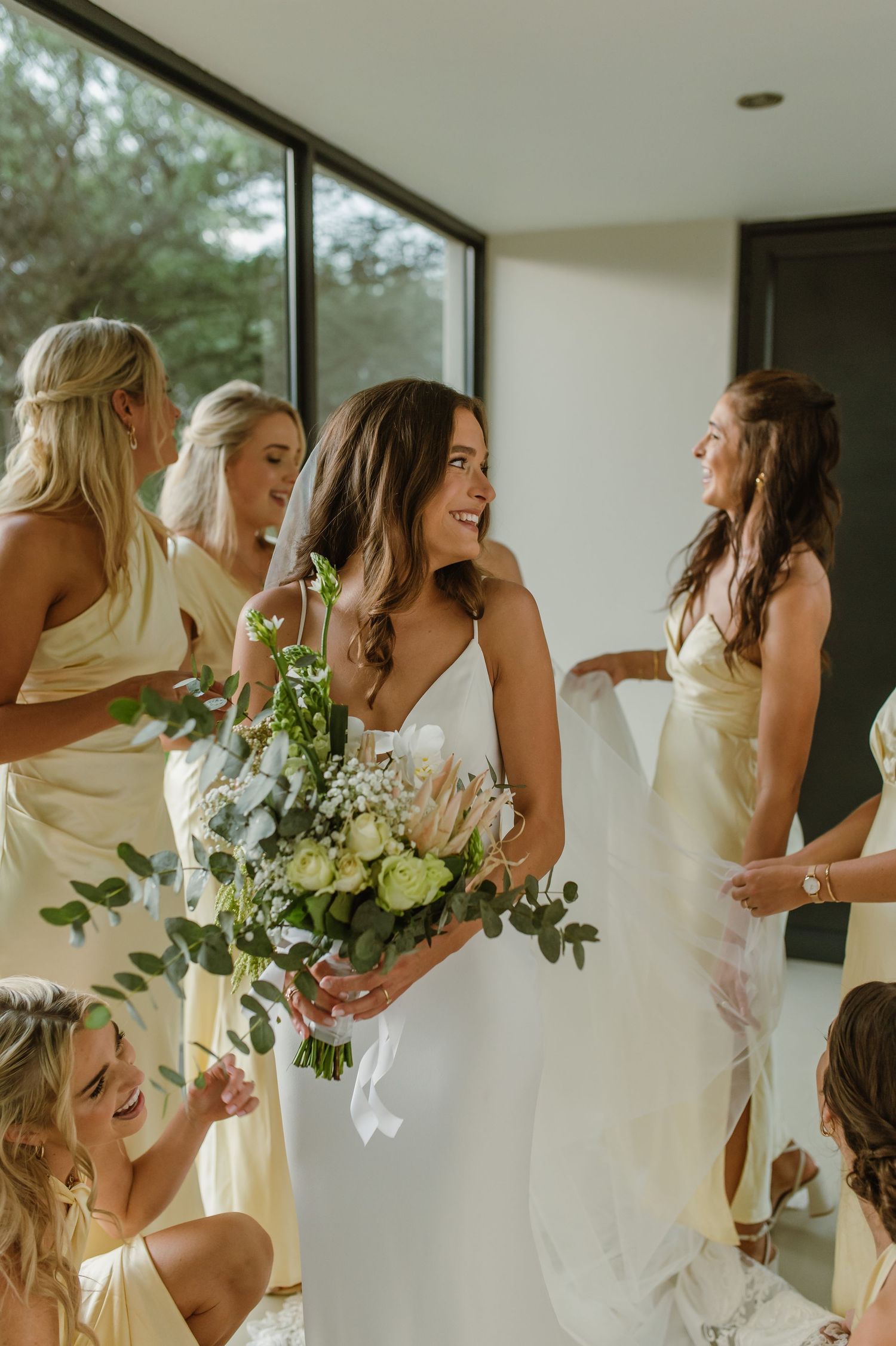 Wedding party in white and yellow dresses sharing candid moments by large windows.