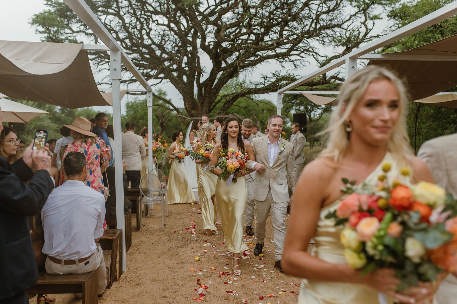 Outdoor wedding ceremony setup with guests seated under shady trees.