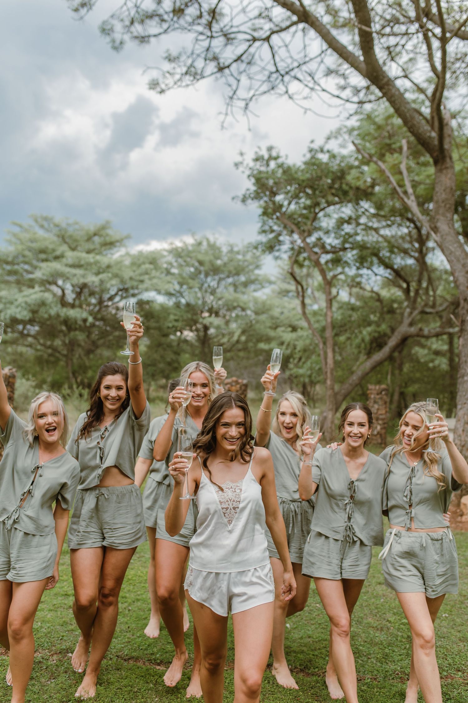Celebration group shot with women in matching outfits raising champagne glasses on lawn.