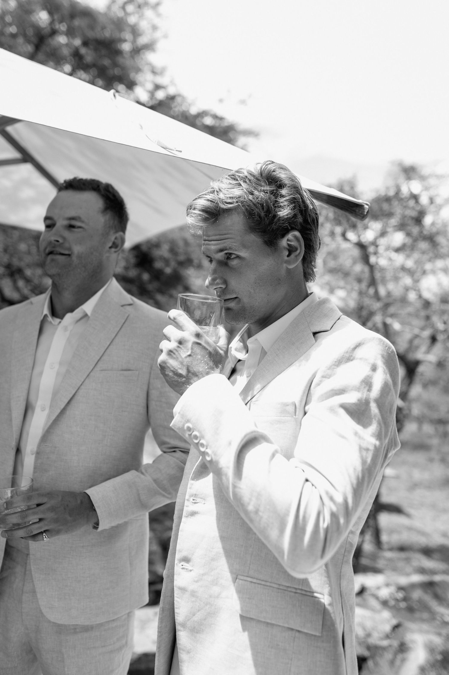 Wedding party members in light suits toast together under a sunny outdoor canopy.