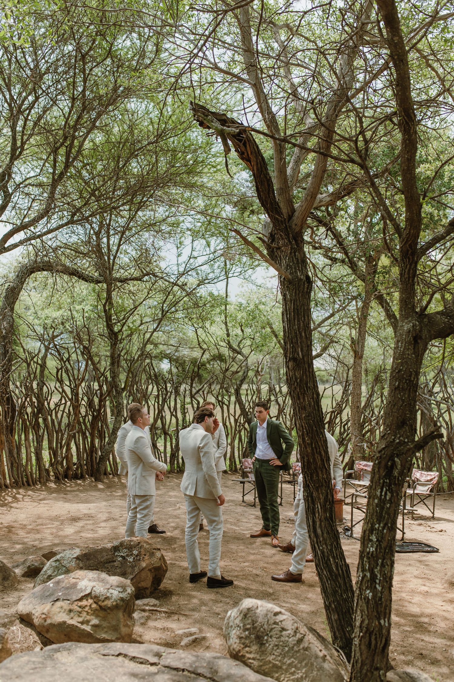 Groomsmen in light colored suits gather in a natural wooded area with scattered rocks.