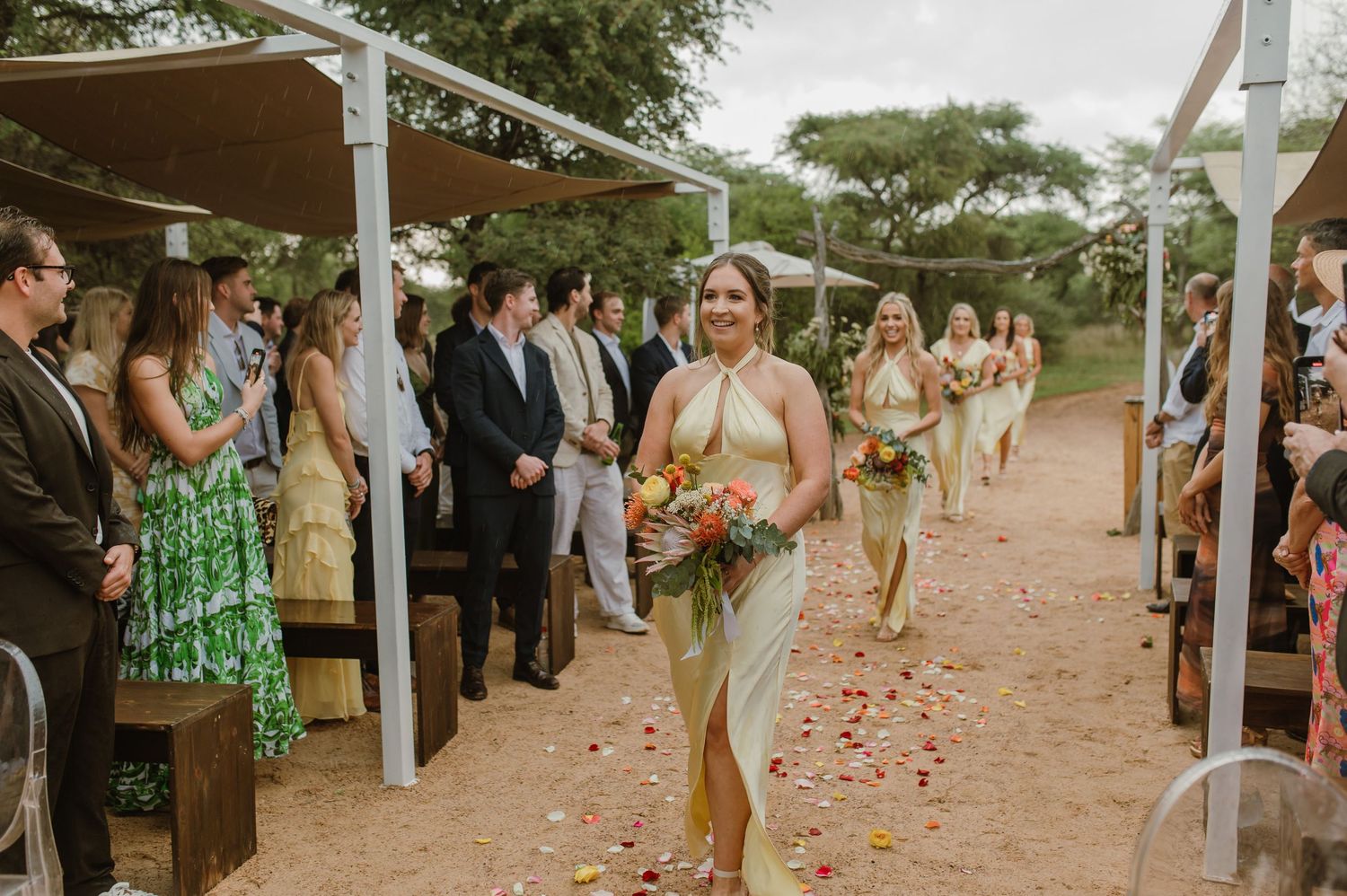 Wedding ceremony processional with guests standing along a petal-strewn aisle under white canopies.