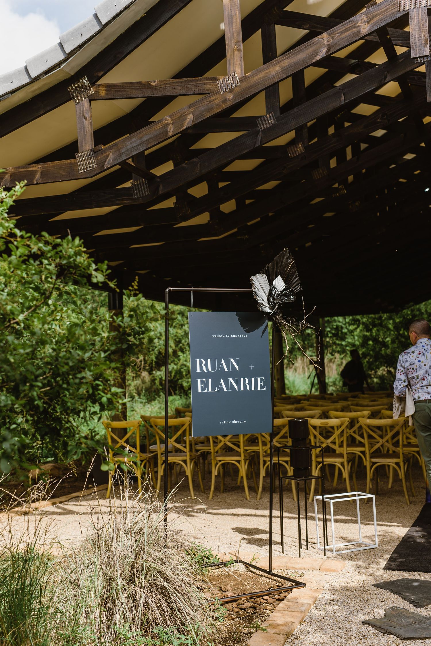Rustic outdoor wedding venue with wooden beams and chairs set up under a covered pavilion in a garden setting.