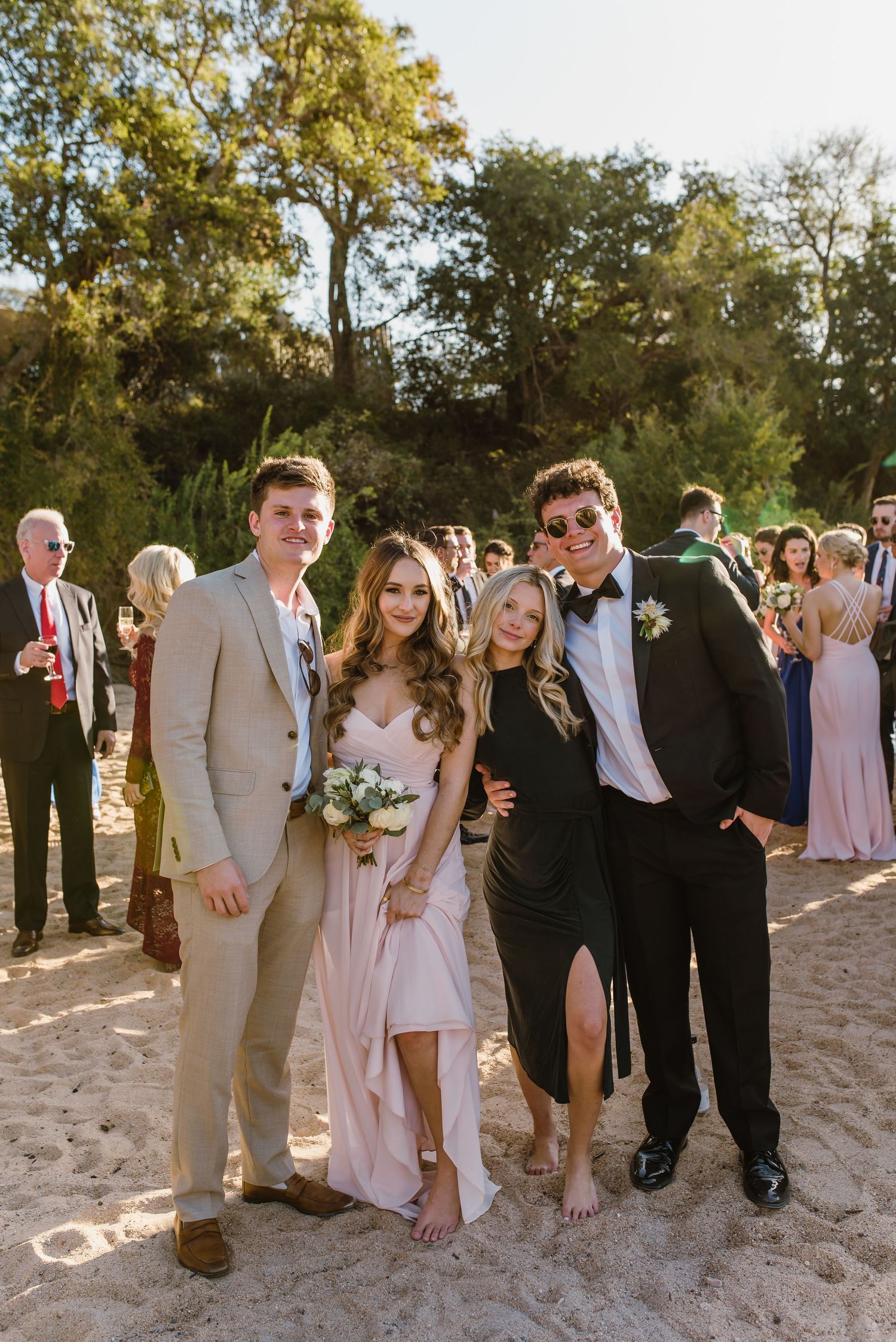 Wedding party poses for group photos on a sandy beach with trees in the background.