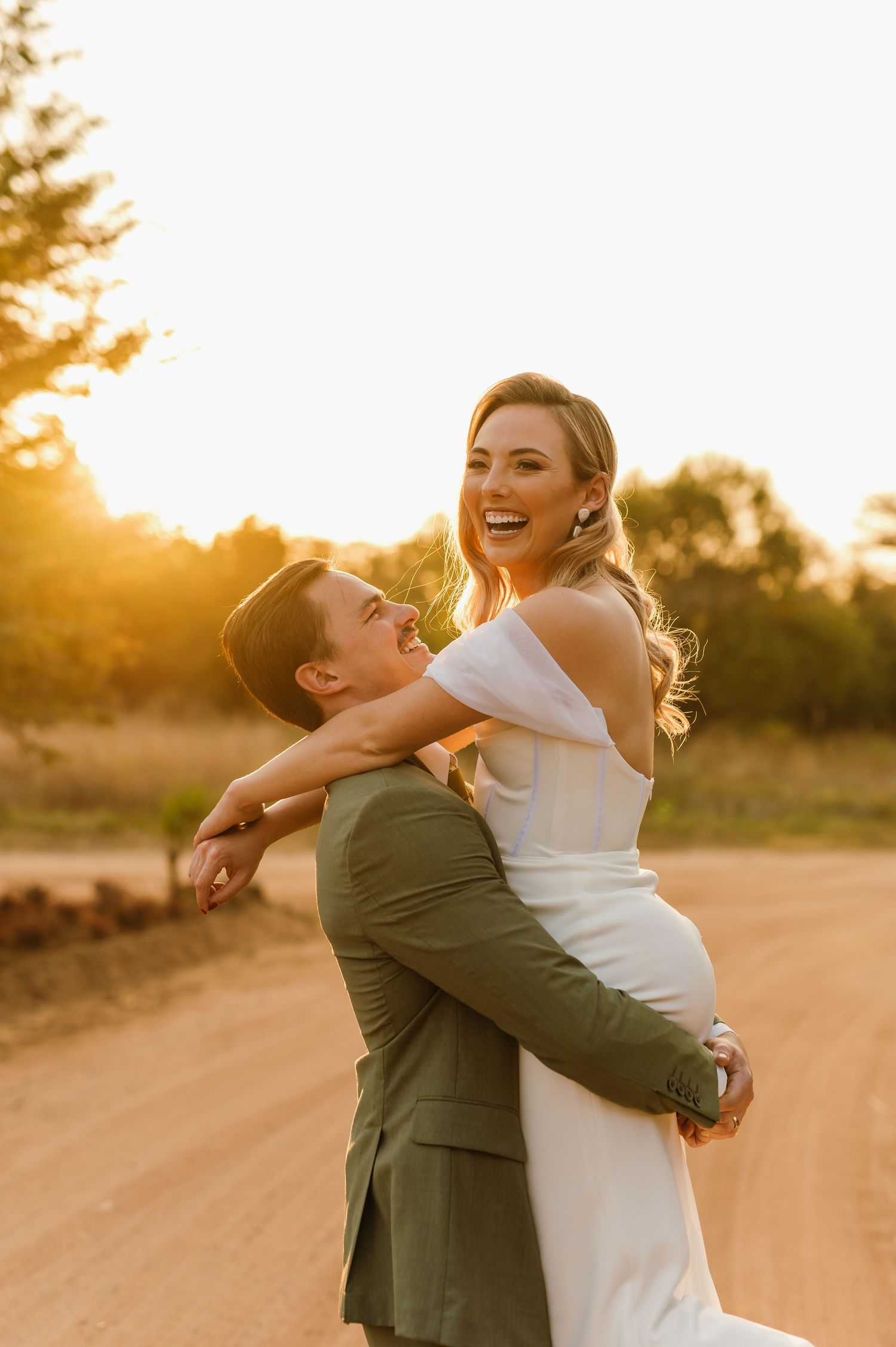 A romantic sunset photoshoot shows a couple embracing joyfully on a dirt road.