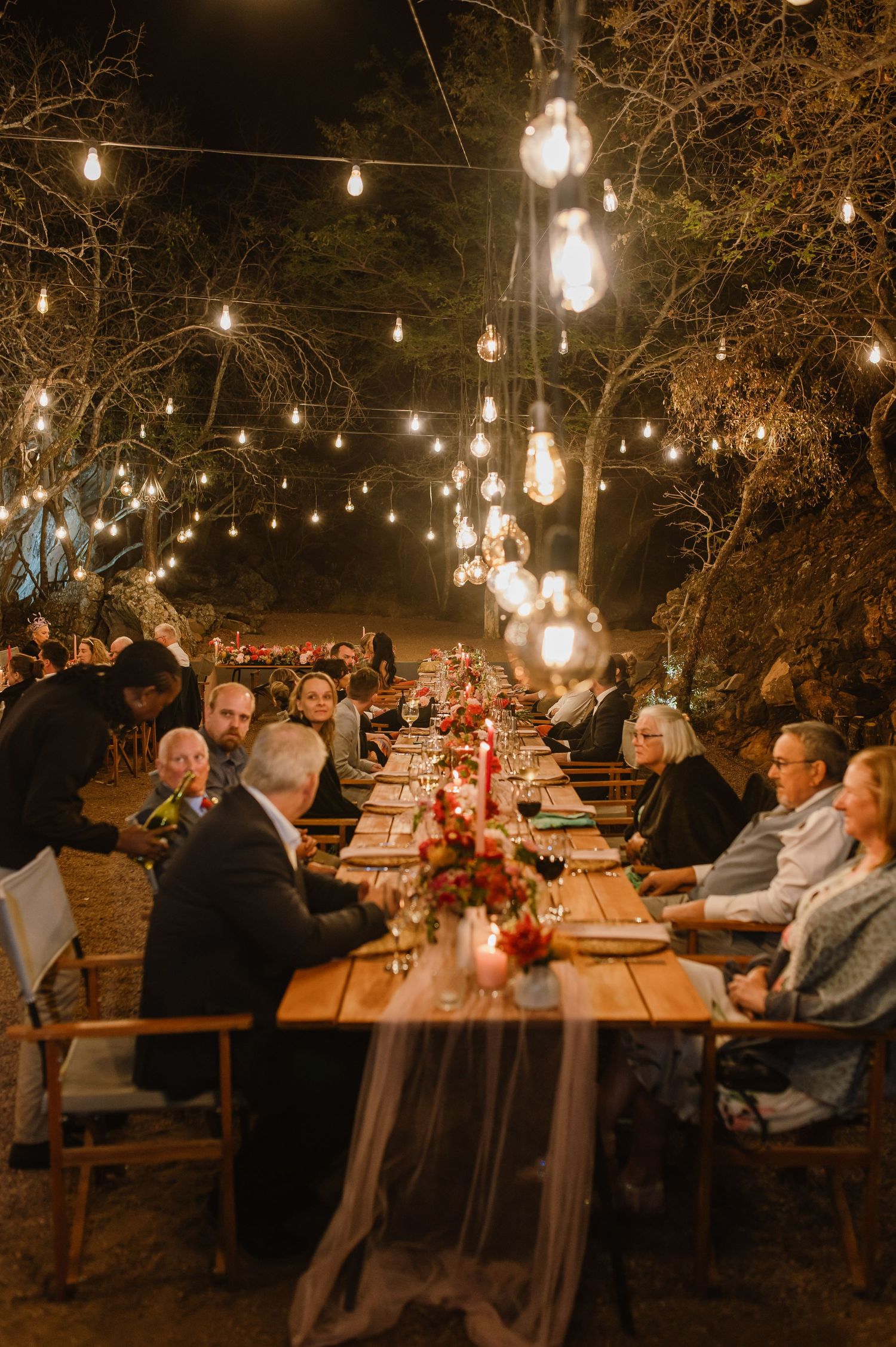 Guests dining at long wooden tables under romantic string lights at an outdoor evening event.