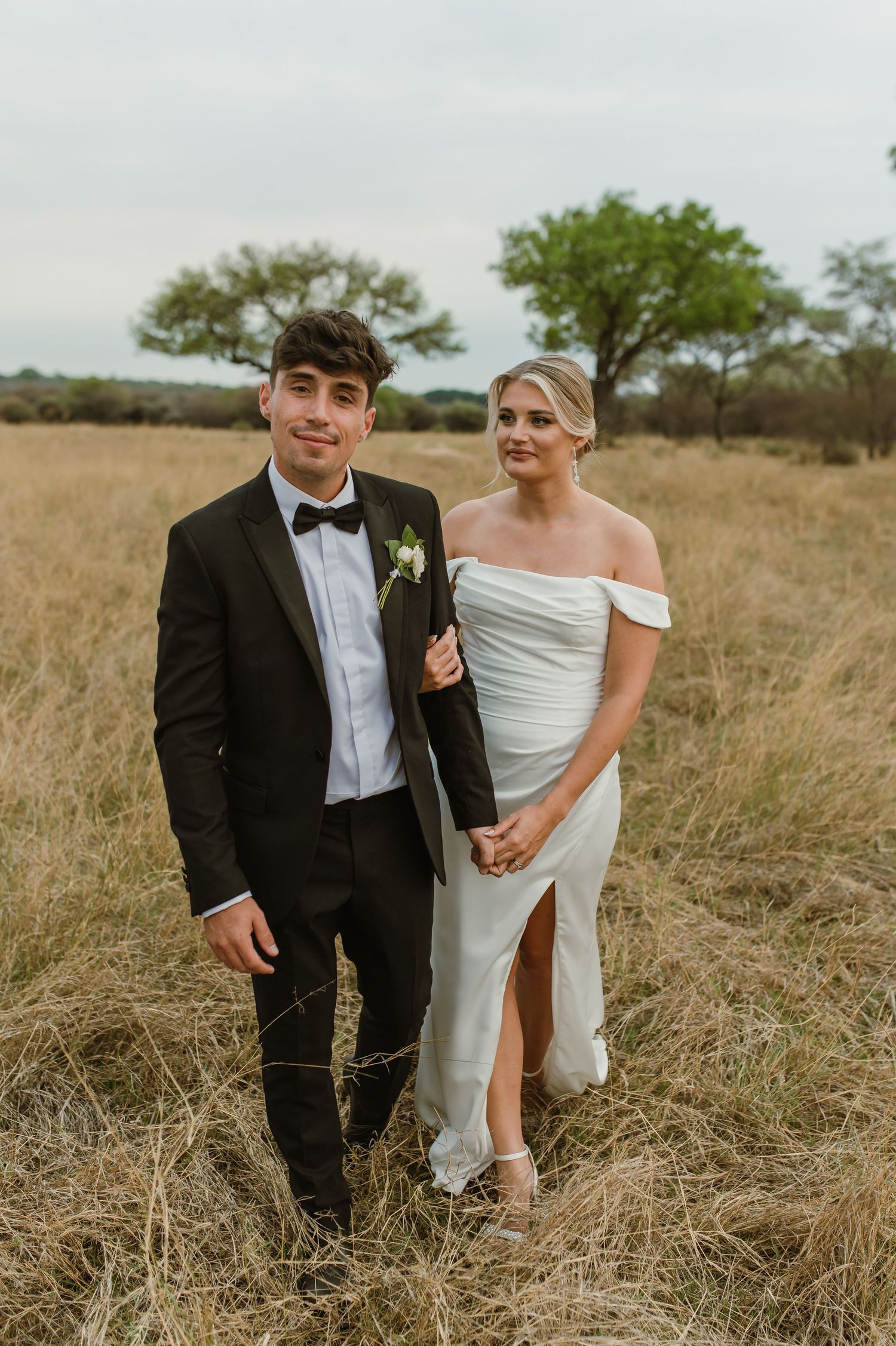 A newlywed couple laughs together while walking through tall grass in a natural setting.