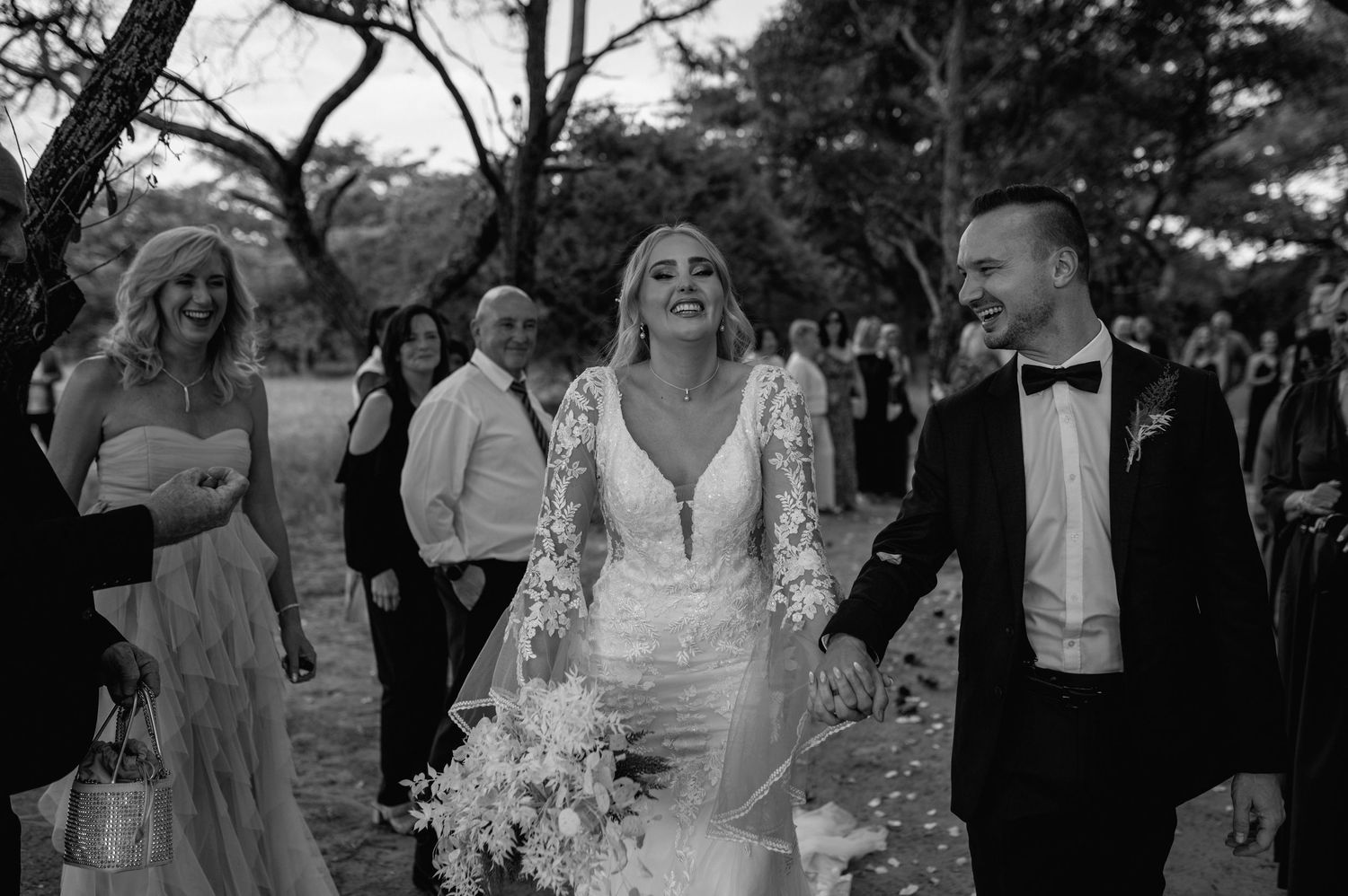 Wedding party celebrates joyfully under trees at an outdoor ceremony.