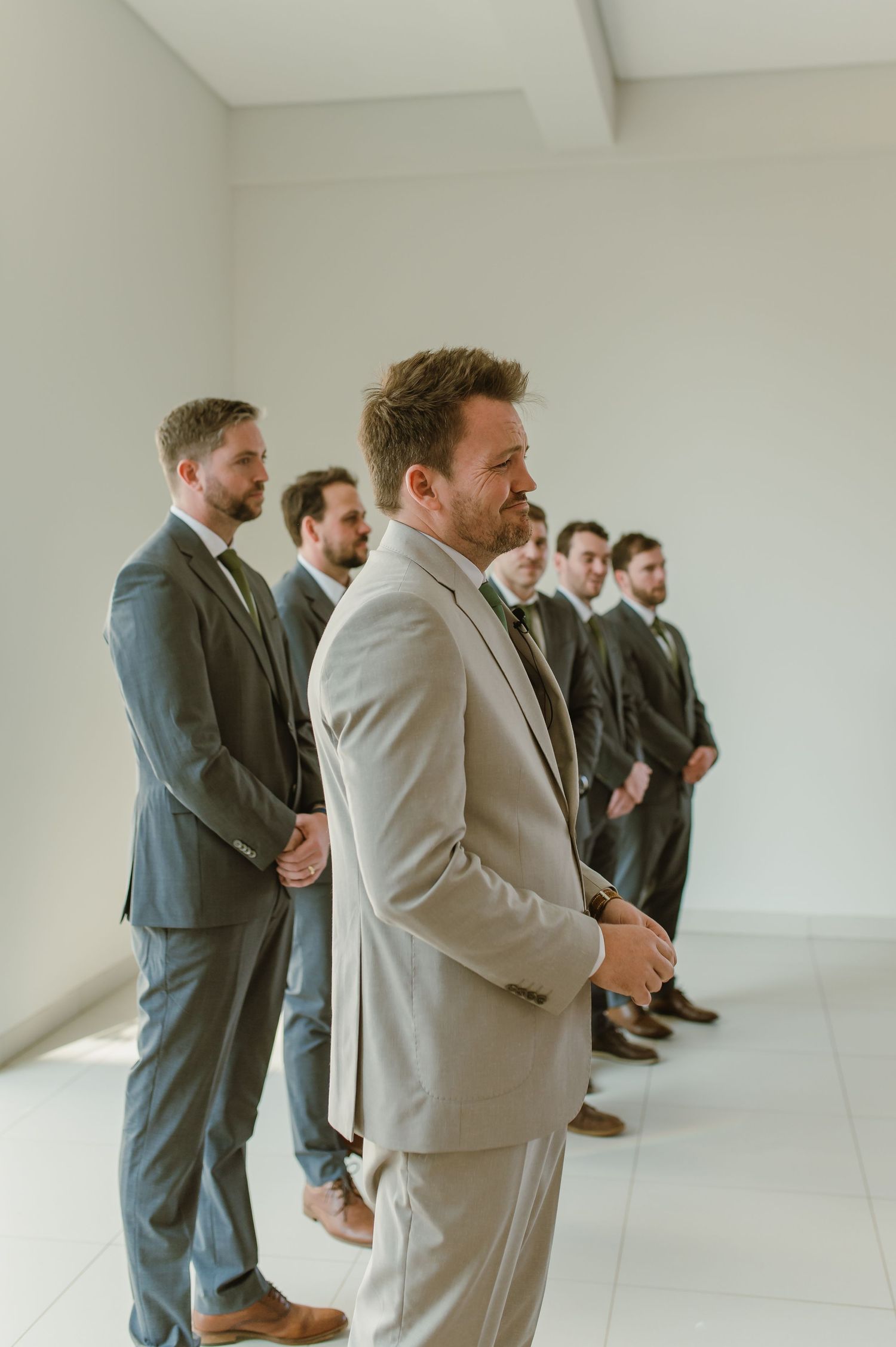 Groomsmen in suits stand in line against white wall during wedding preparations.
