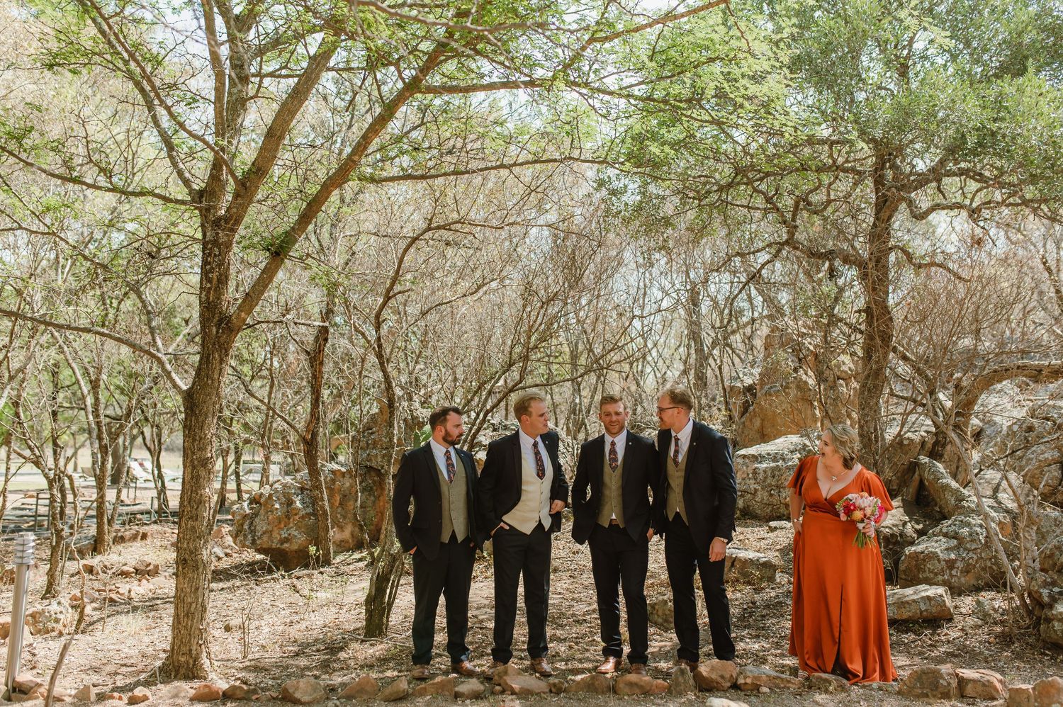 A wedding party stands together on sandy ground surrounded by bare winter trees.