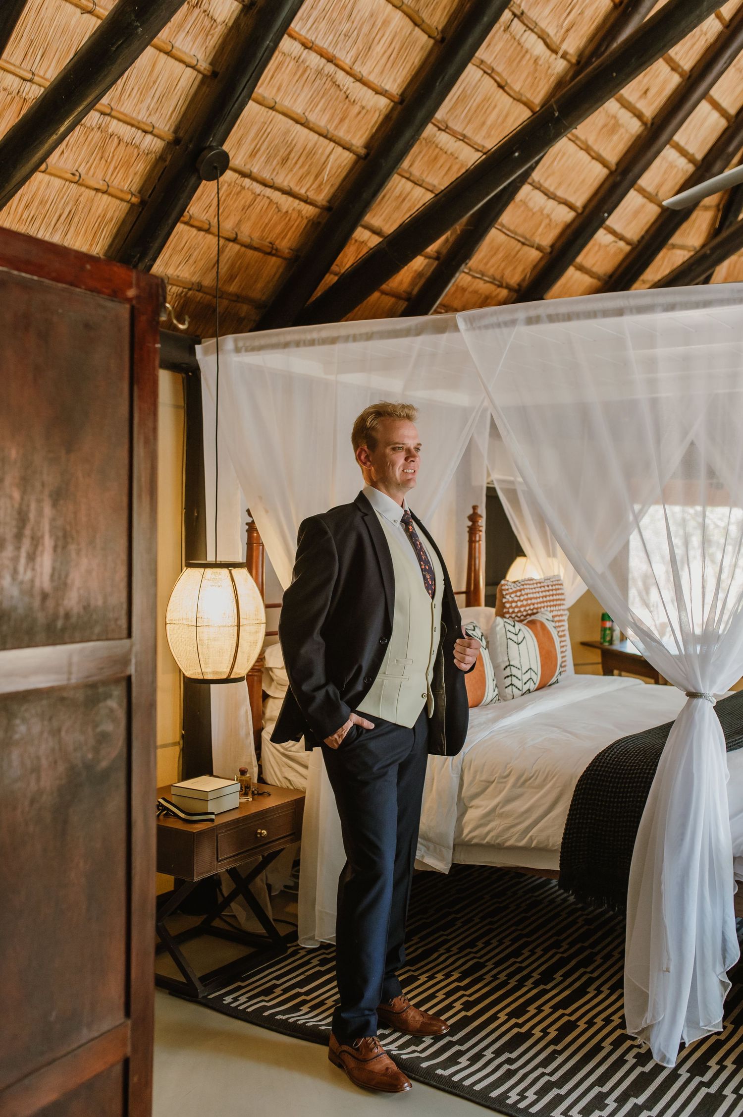 Person in a suit and vest poses in an elegant bedroom with lantern lighting and mosquito netting.