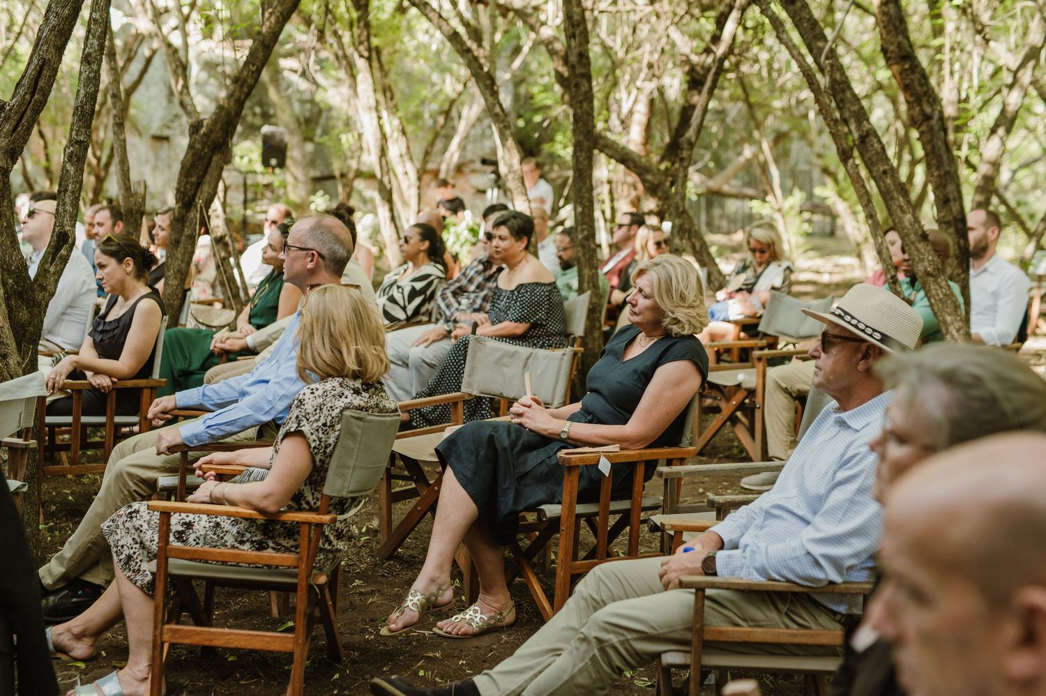 Wedding guests seated on wooden benches under forest canopy for outdoor ceremony.