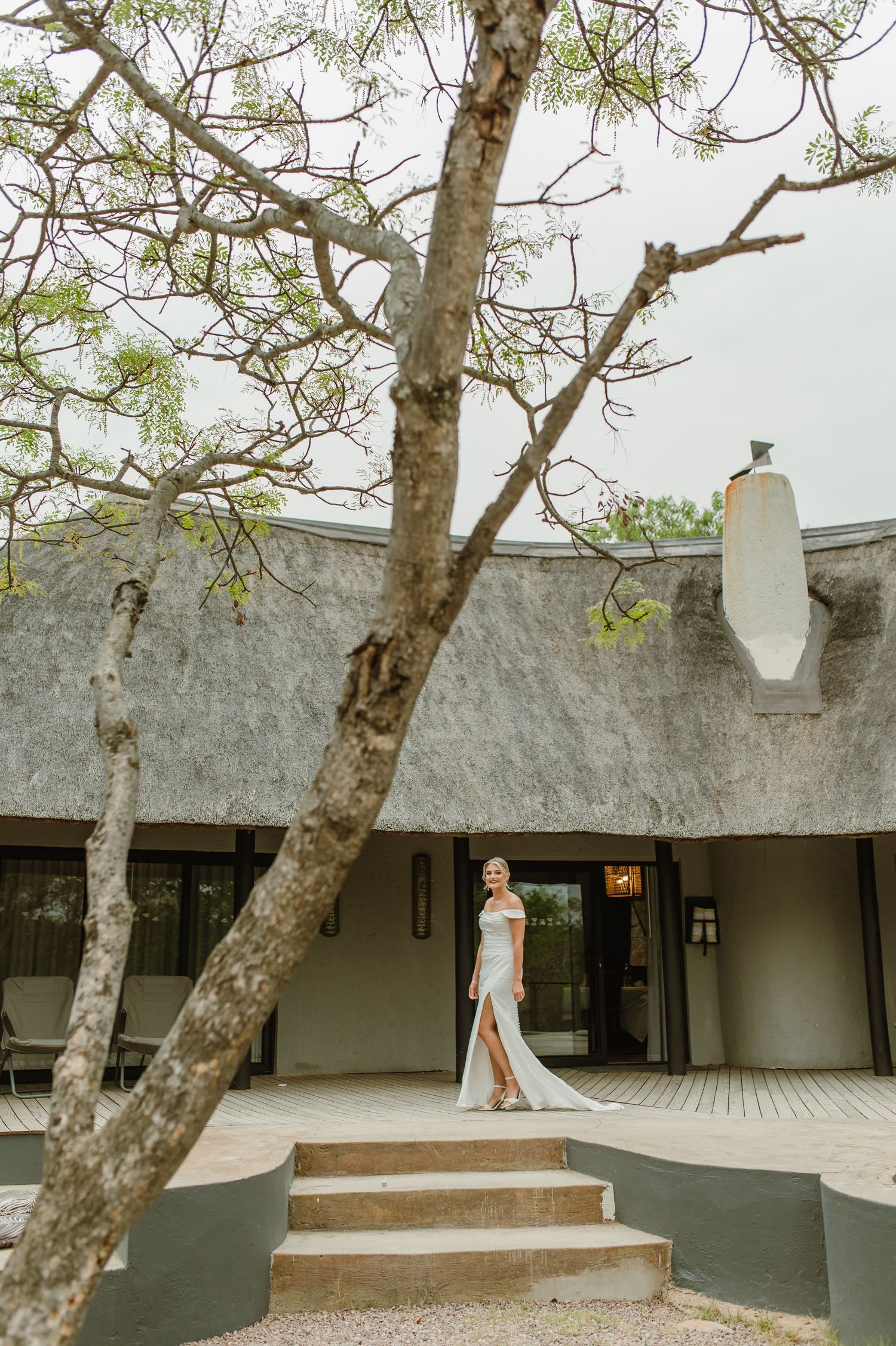 Bride in white gown stands on steps of thatched-roof building surrounded by trees.