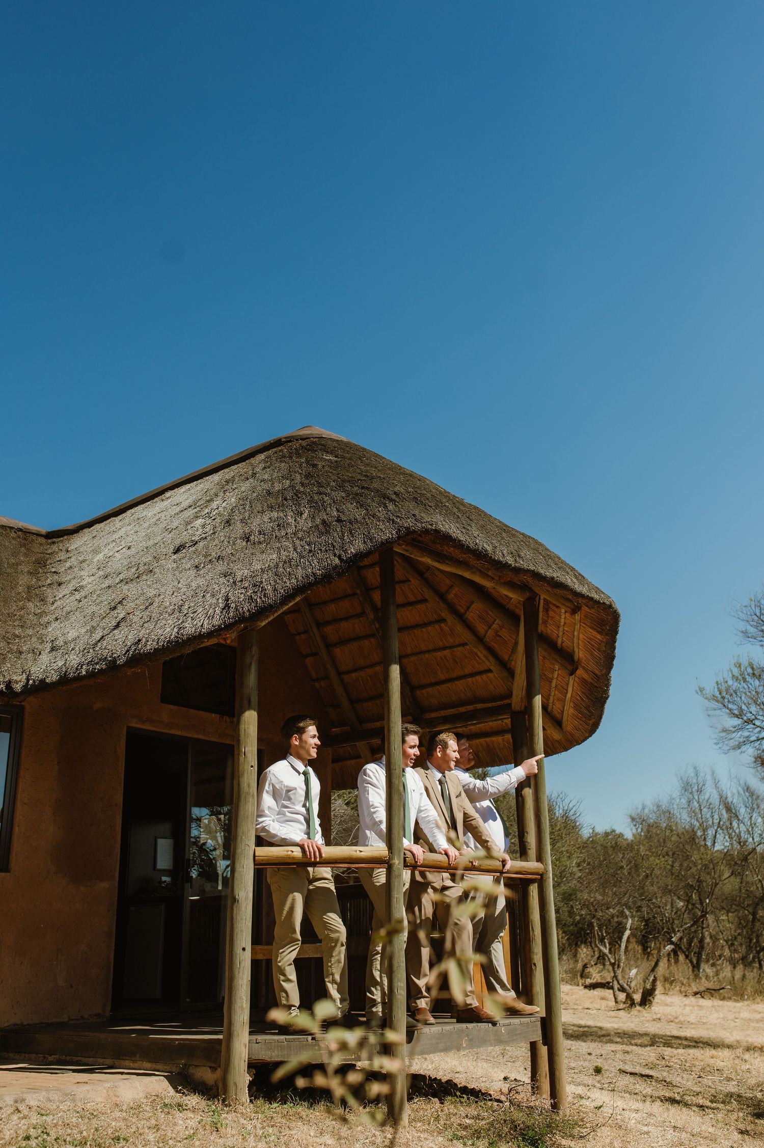 Group relaxing outside traditional African lodge with thatched roof against blue sky.