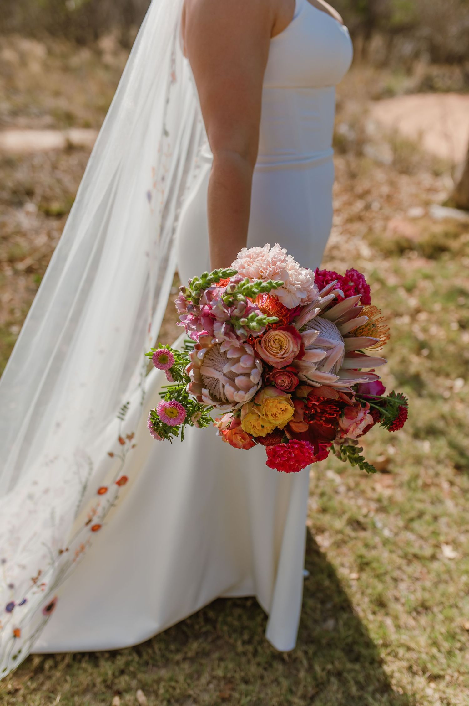 Vibrant bridal bouquet with red, pink and orange flowers against a white wedding dress.