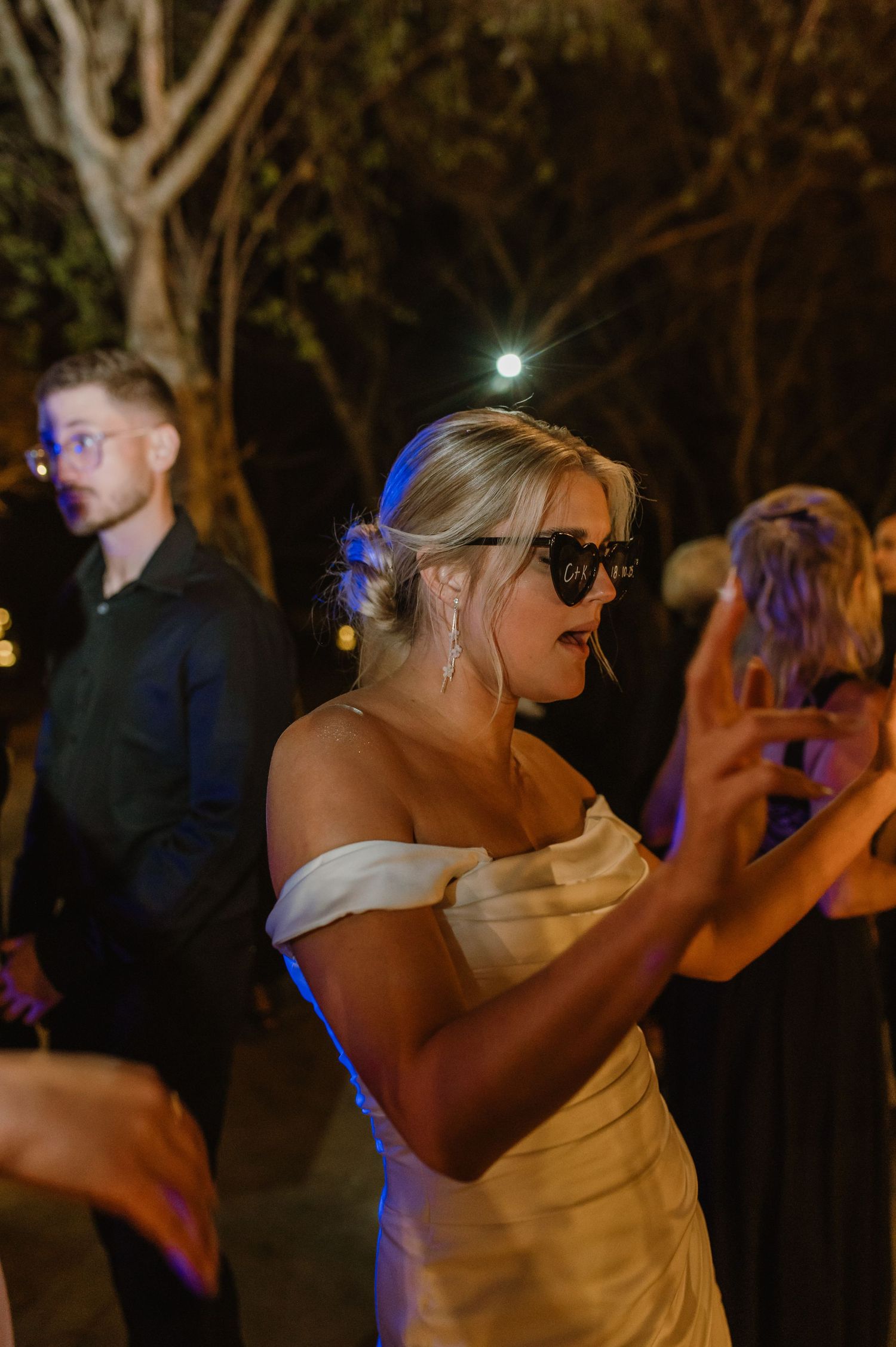 People dancing under trees at a nighttime outdoor wedding reception with blue accent lighting.