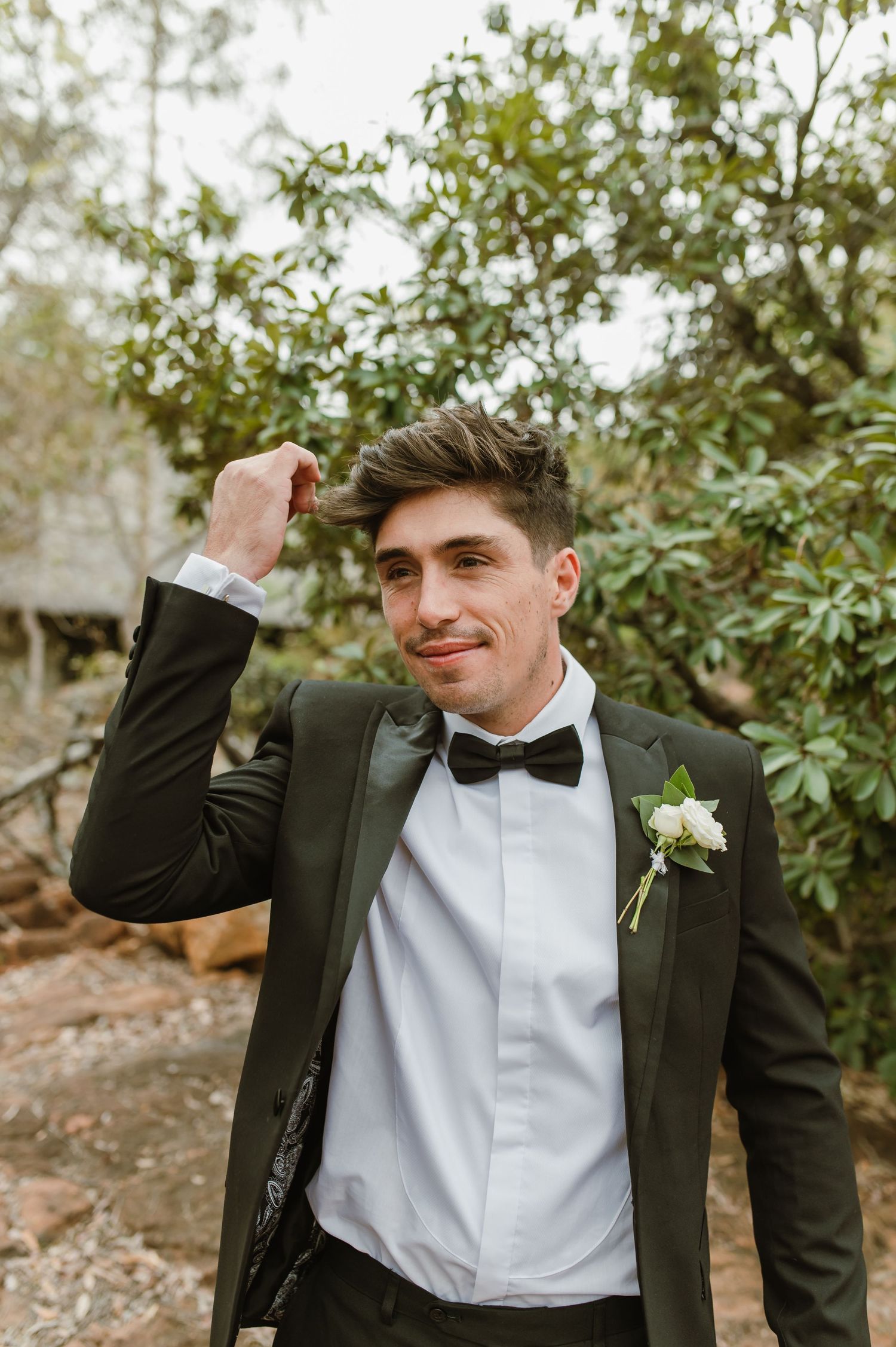 Groomsman in black tuxedo and bow tie making animated gestures outdoors.