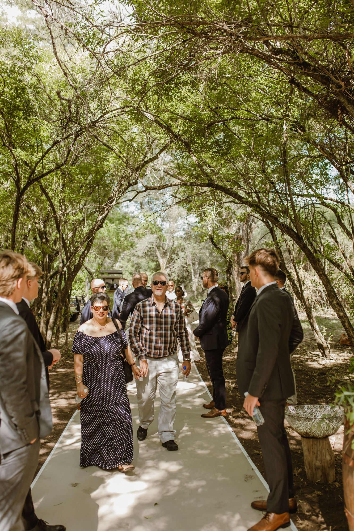 Wedding attendees mingle on a shaded pathway beneath overhanging trees.