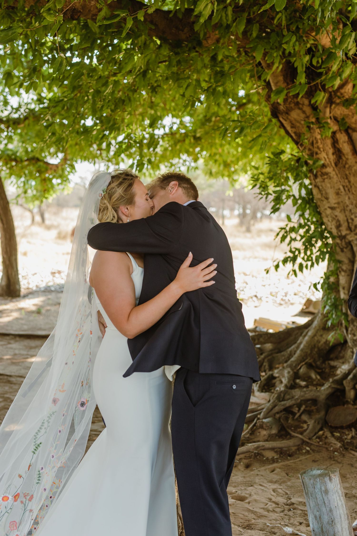 Newlyweds share their first kiss under a canopy of green leaves.
