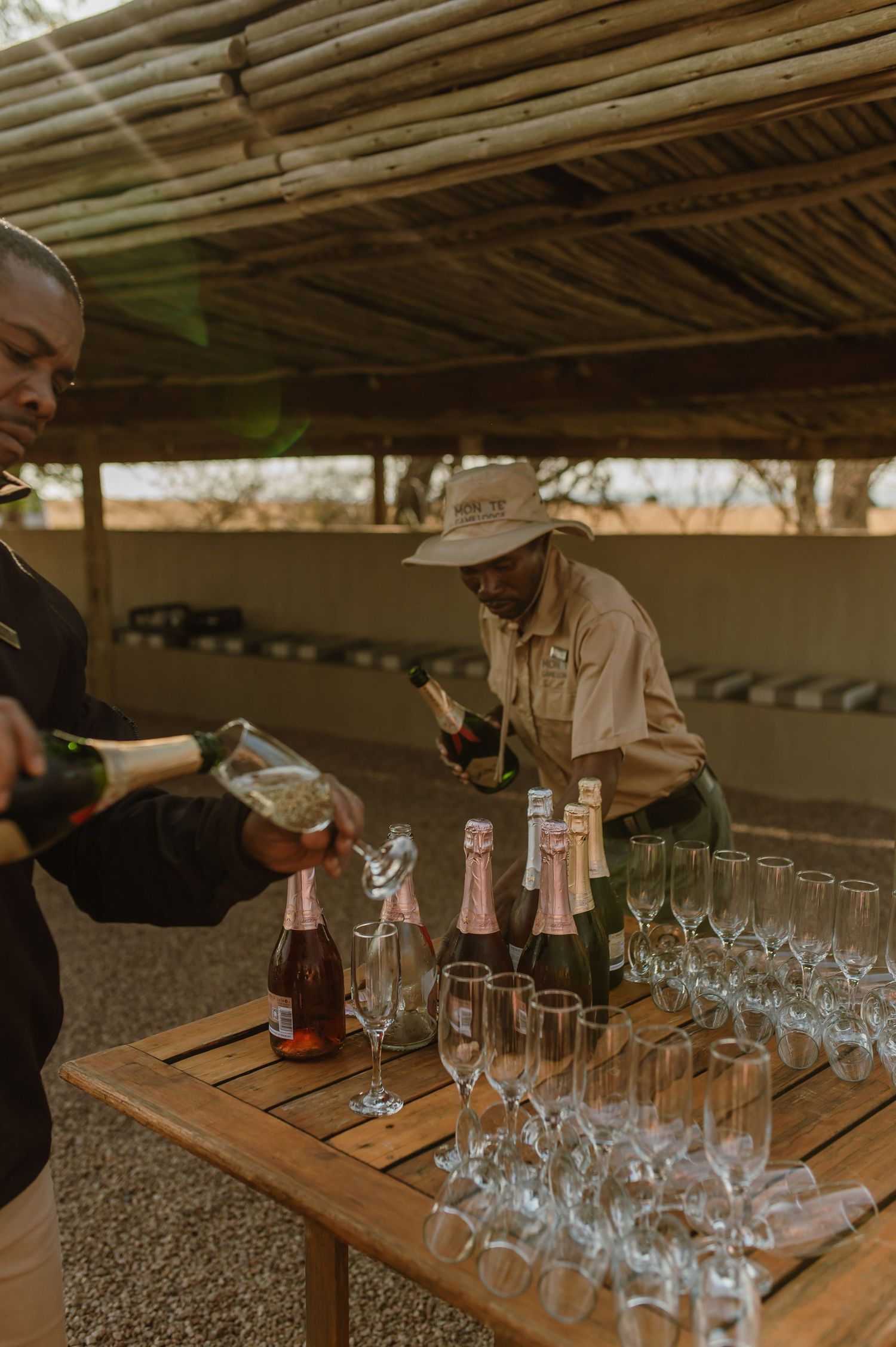 Bar setup with rows of empty wine glasses on wooden tables under rustic shelter.