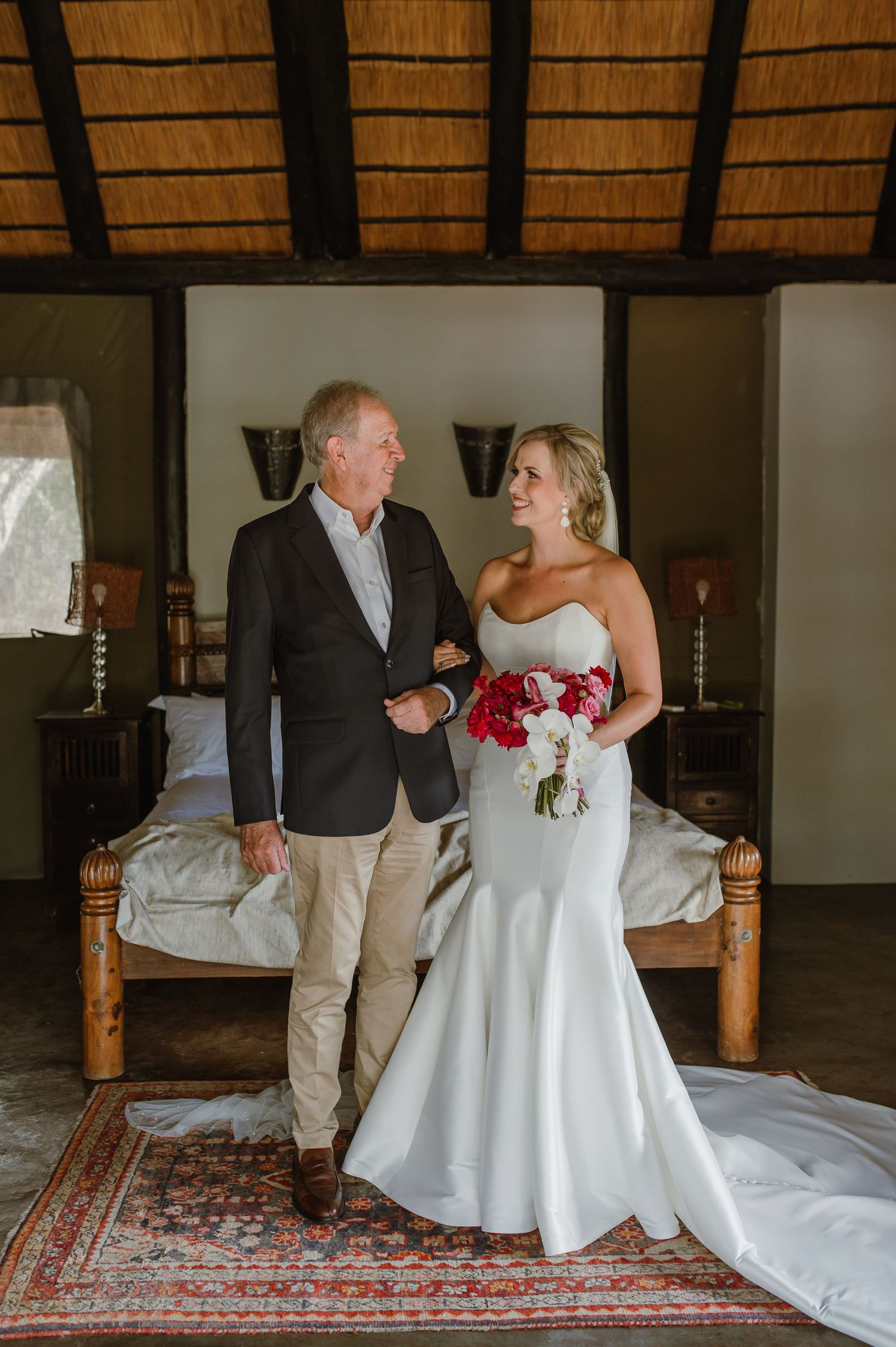 Sweet moment between two people in formal attire on a bed with thatched roof overhead.