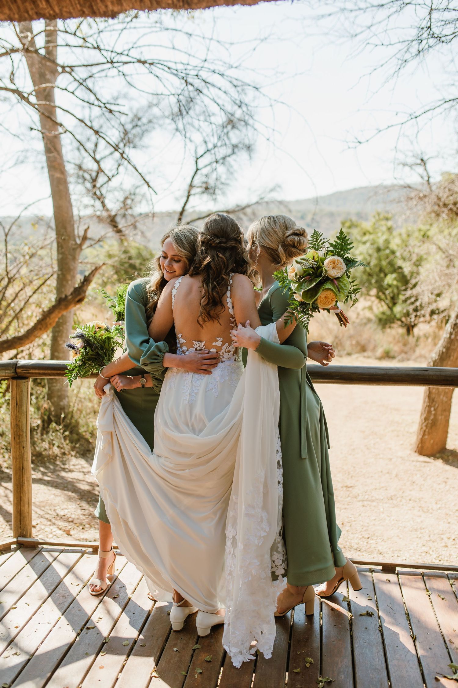 Bridal party in green dresses helping with wedding dress on rustic wooden deck.