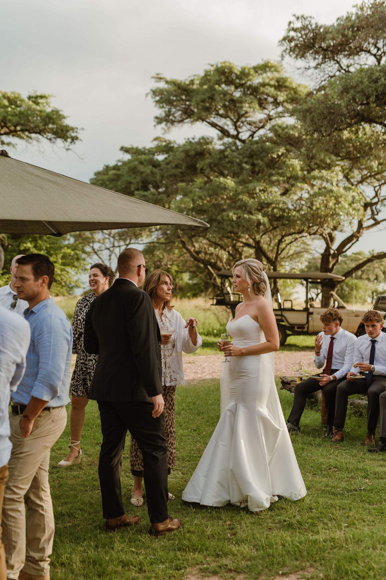 Wedding guests gathered on a grassy lawn under acacia trees during an outdoor celebration.