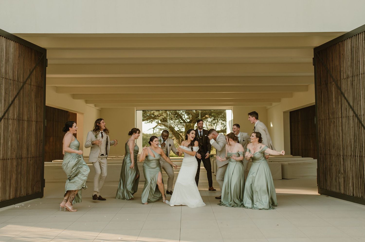 Wedding party gathers in covered walkway wearing sage green and neutral formal attire.