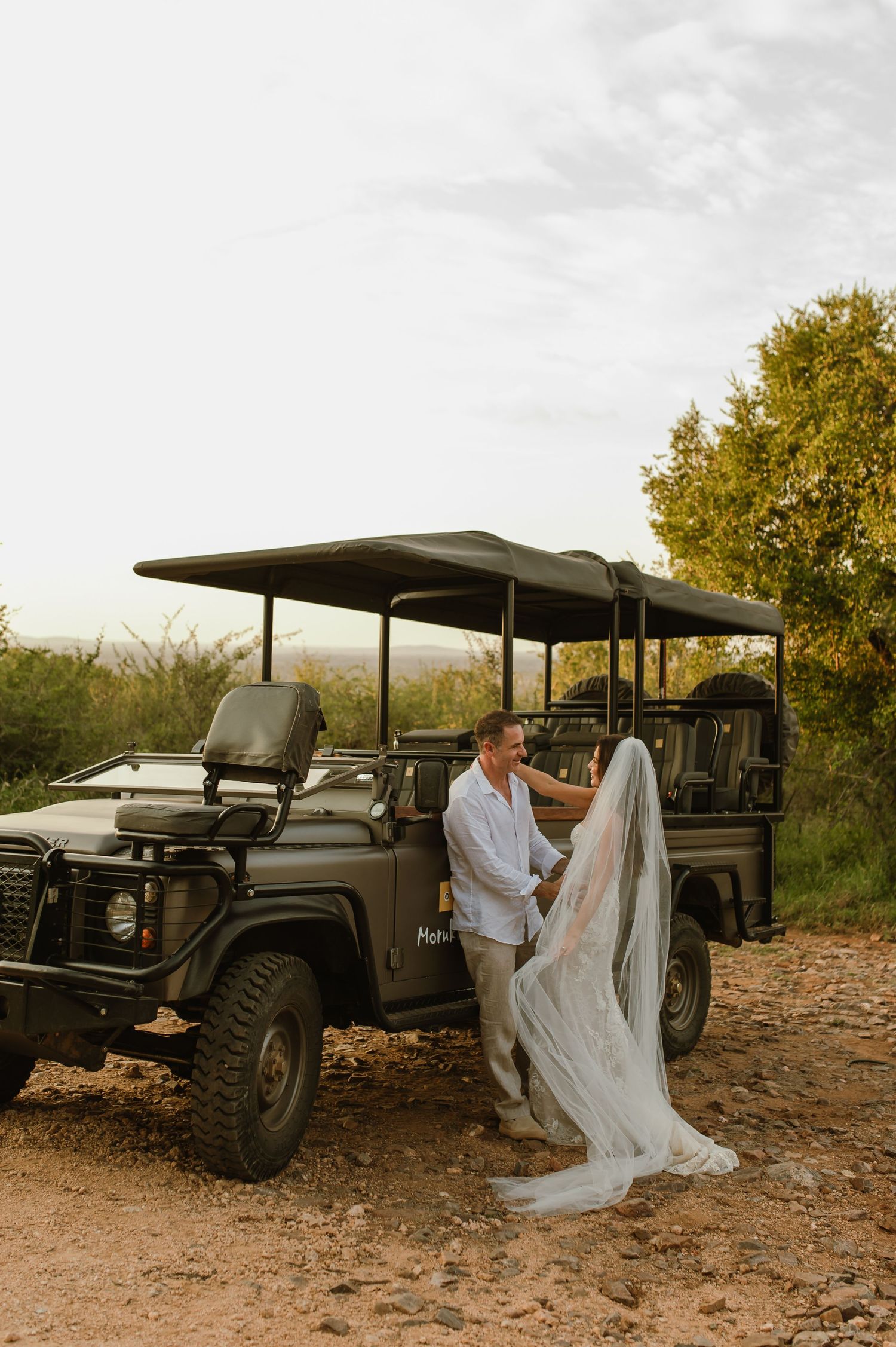 Bride and groom holding hands beside green safari Land Rover on dusty trail at wedding.