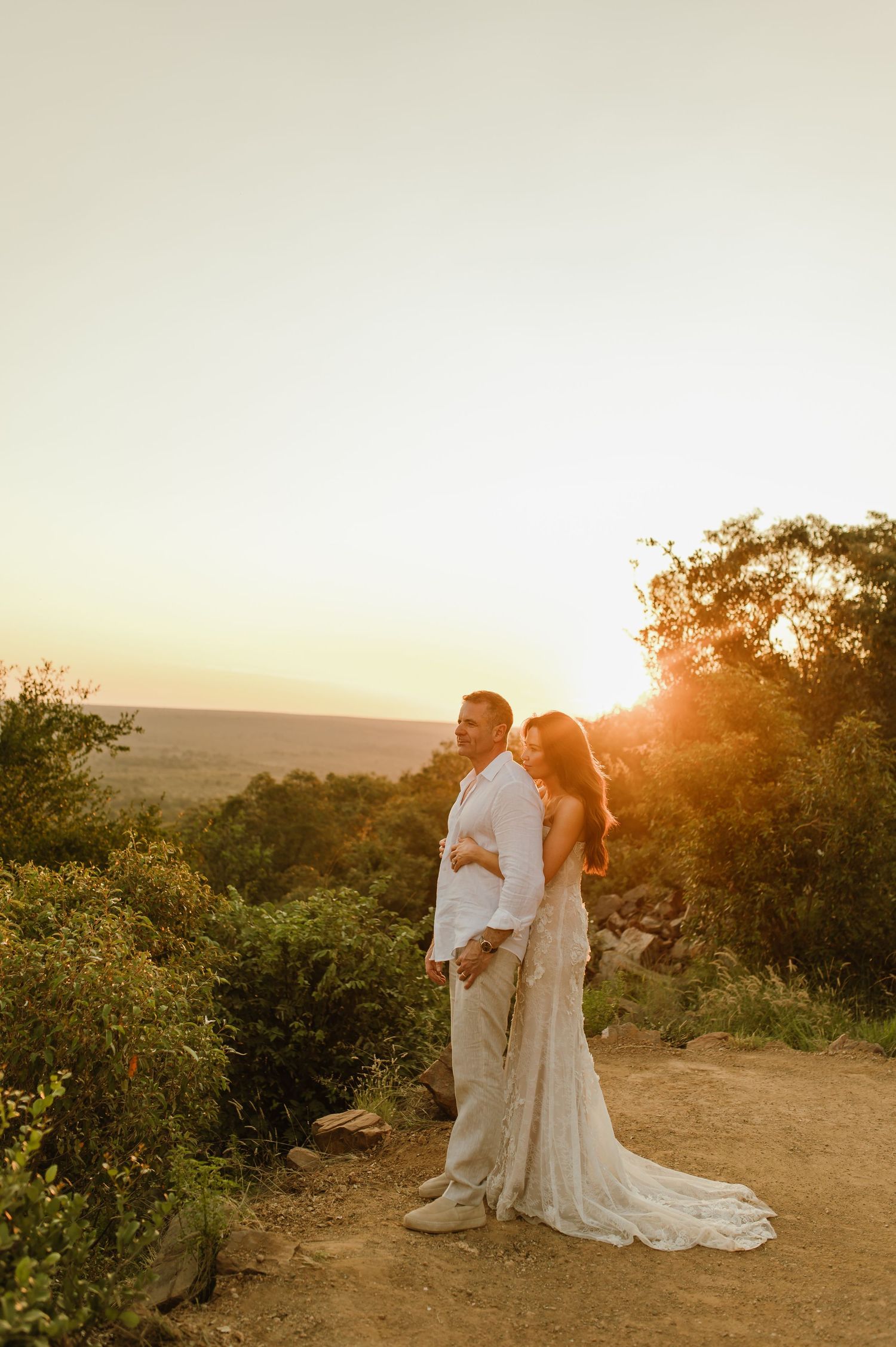 Couple embraces at golden sunset on a scenic overlook, bride in lace gown and groom in white shirt.