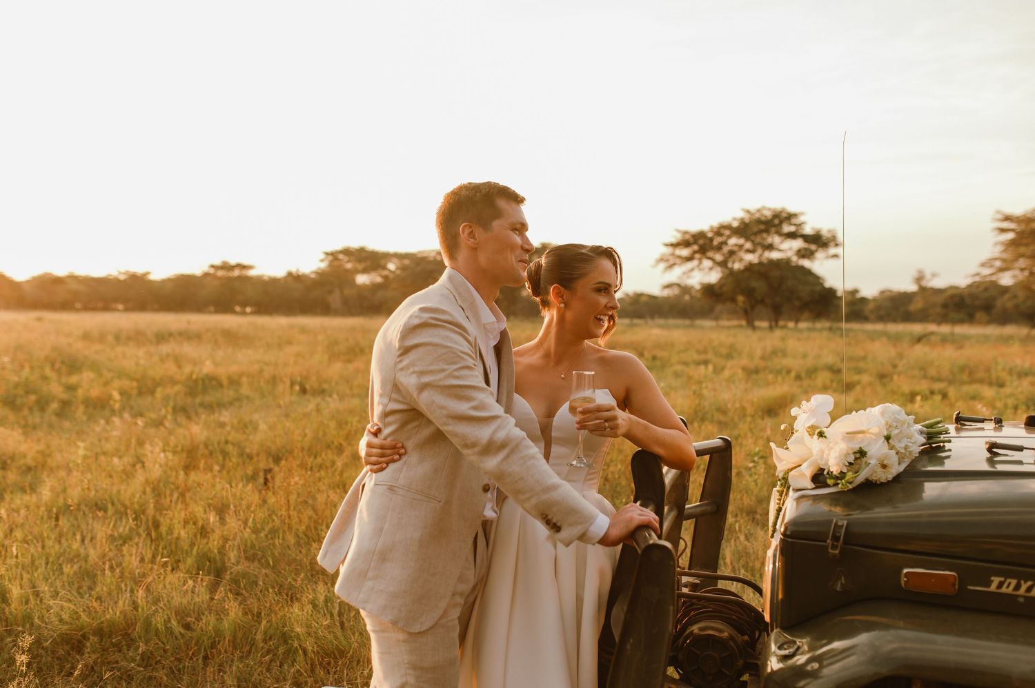 A couple shares intimate moments near a vintage car in a golden-lit field at sunset.