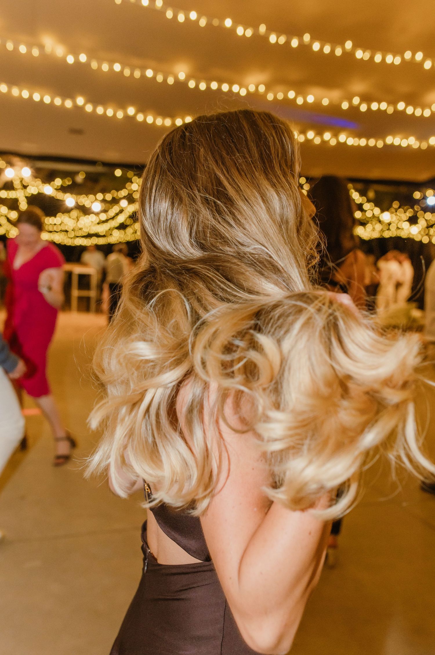 Blonde hair swirls in motion on a dance floor under warm string lights at an indoor event venue.
