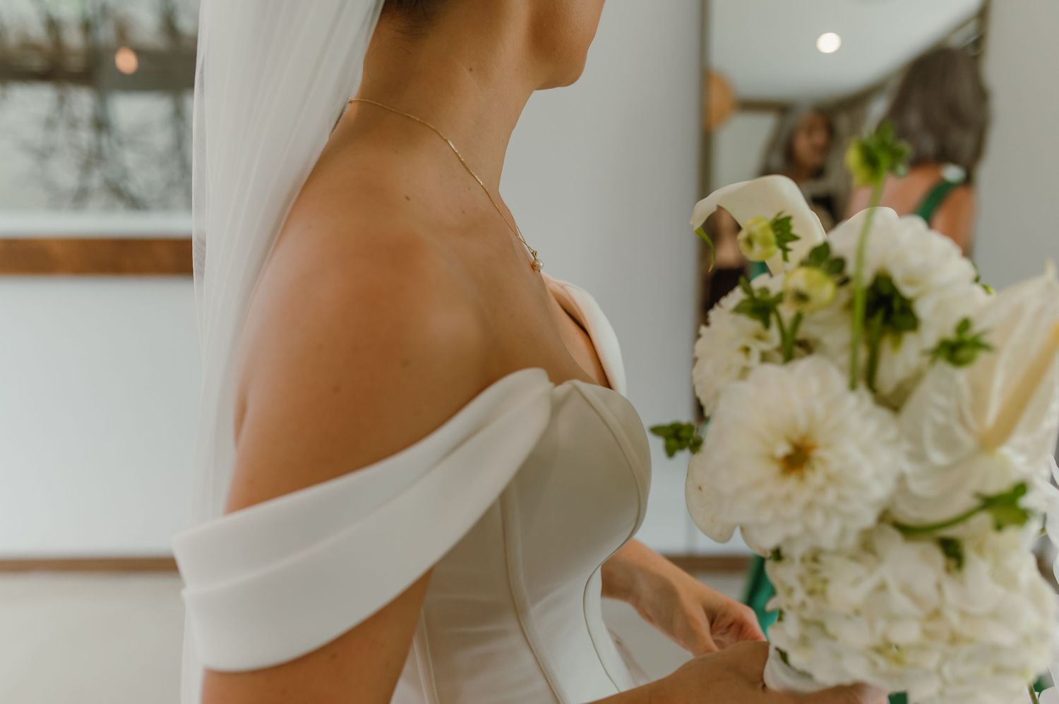 Close-up detail of an elegant off-shoulder white wedding dress with white floral arrangements in the background.