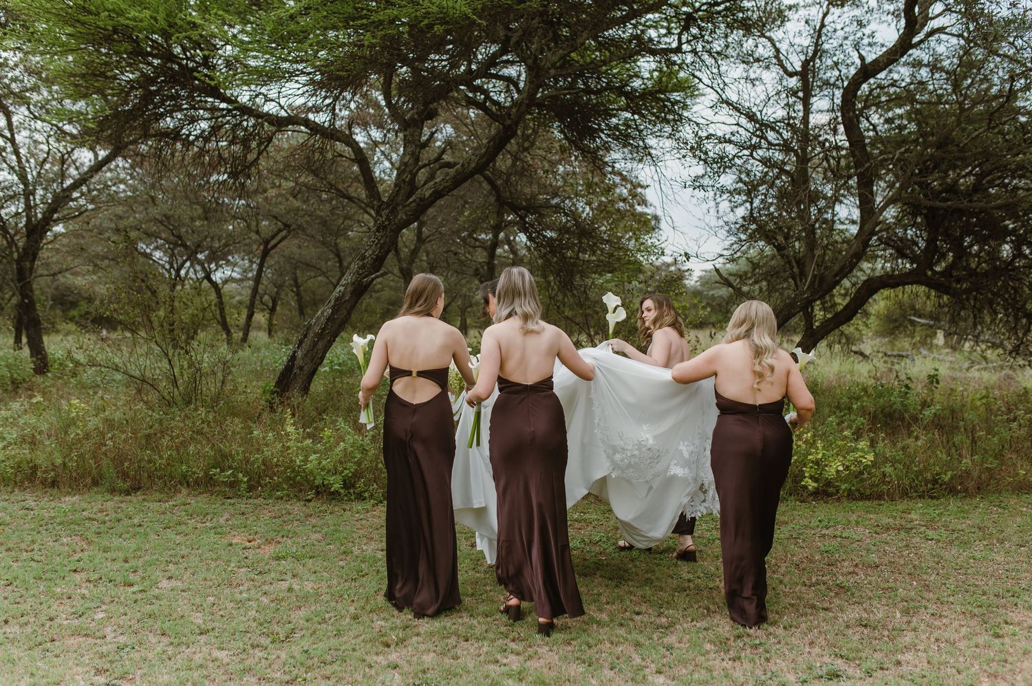 Bridesmaids in brown dresses help carry wedding dress train through outdoor garden setting.